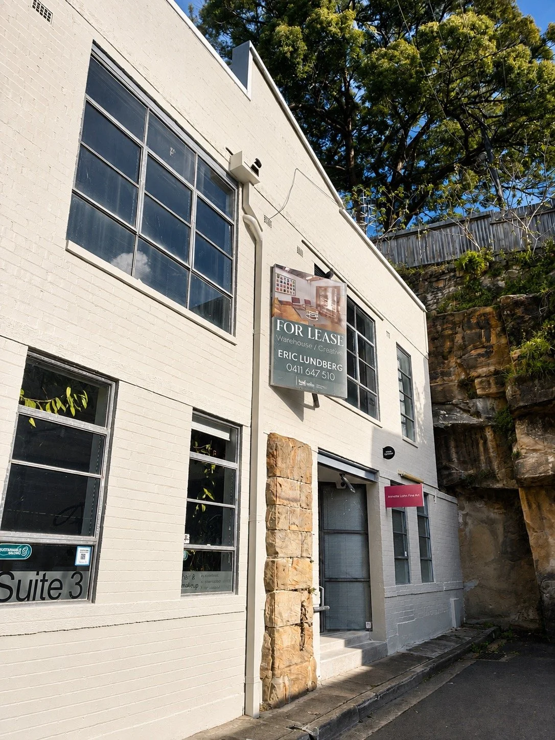 A gallery in Paddington called Roslyn Oxley9 - white brick building with large windows on a sunny day, with signs indicating a space for lease and an arts room, neighboring natural rocky formations and trees.