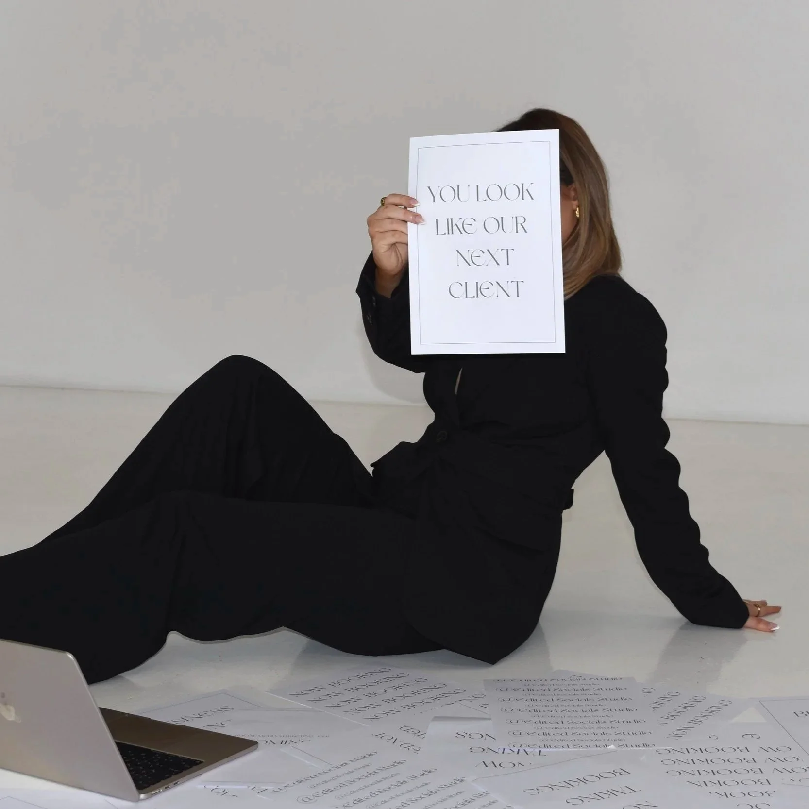 Woman in black suit sitting on the floor, holding a sign that reads 'You look like our next client', with papers scattered around and a laptop open in front.