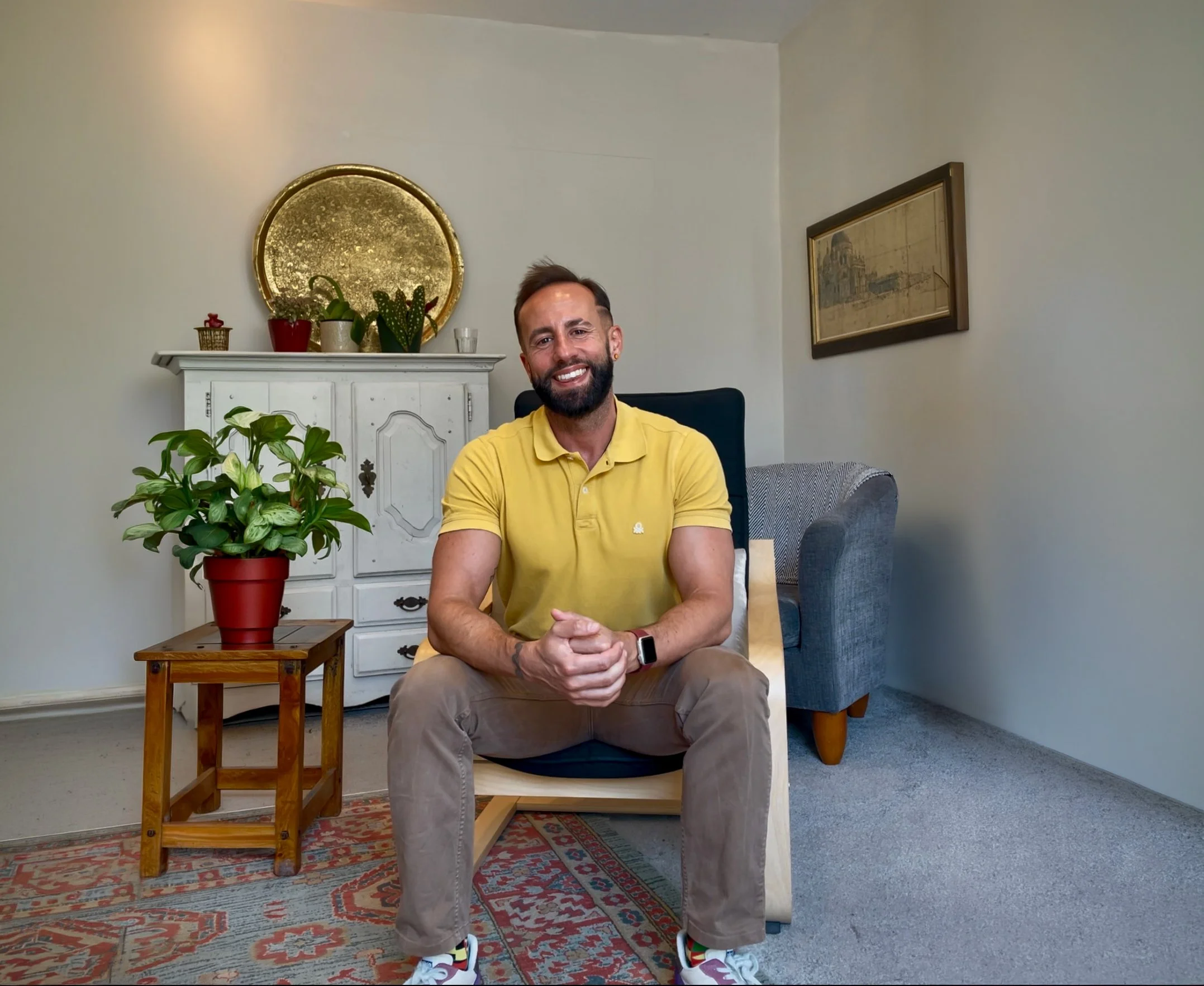 A man sitting on a wooden stool in a living room smiling at the camera, wearing a yellow shirt, beige pants, and colorful sneakers. There are houseplants and decorative items behind him, including a white cabinet, a round gold mirror, and framed artwork on the wall.