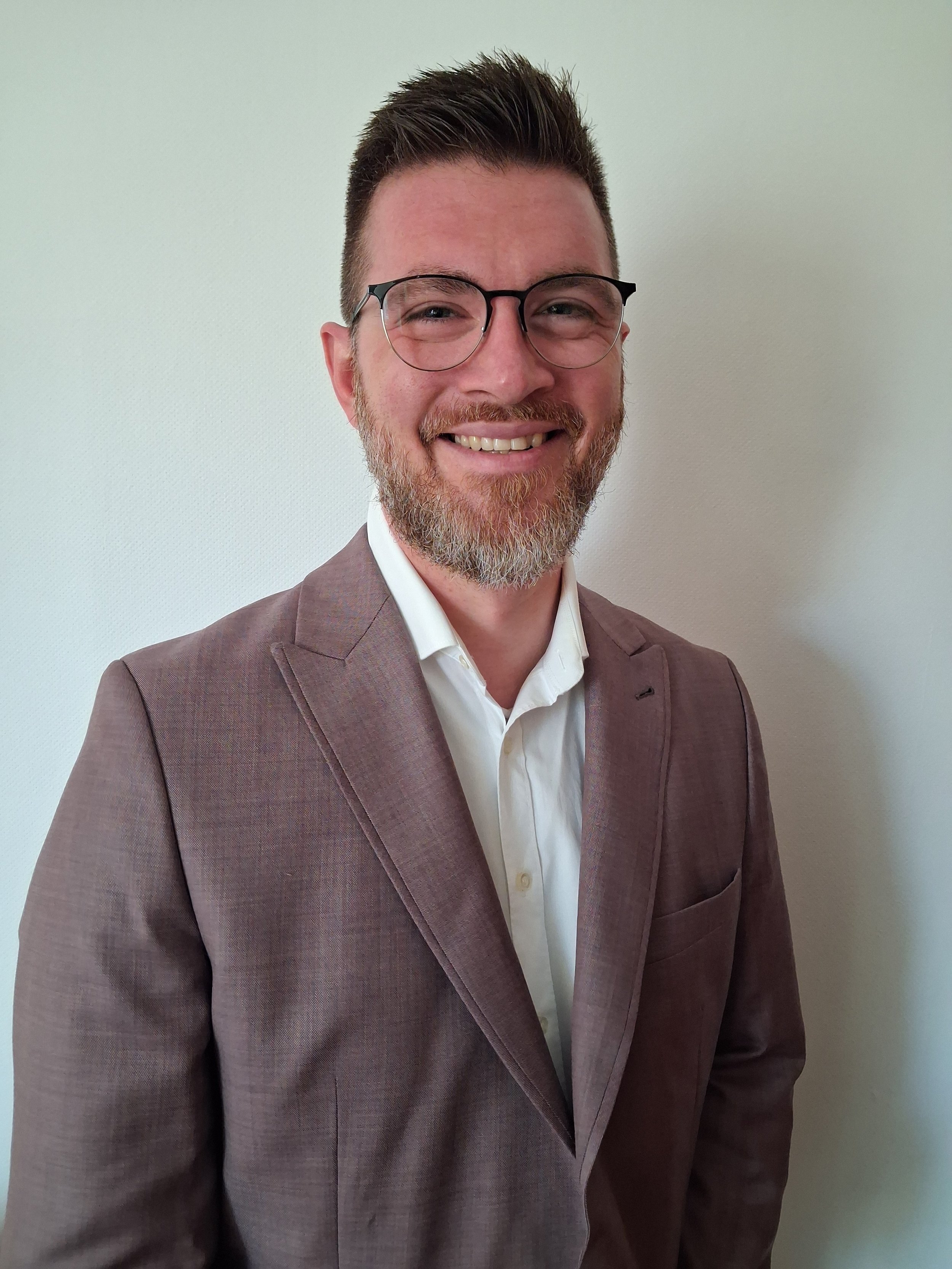 Ein lachender Mann mit Brille in Business-Kleidung vor einer weißen Wand.