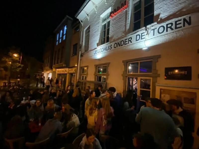 Nighttime scene outside a building with a sign in Dutch and a crowd of people gathered, some sitting and some standing.