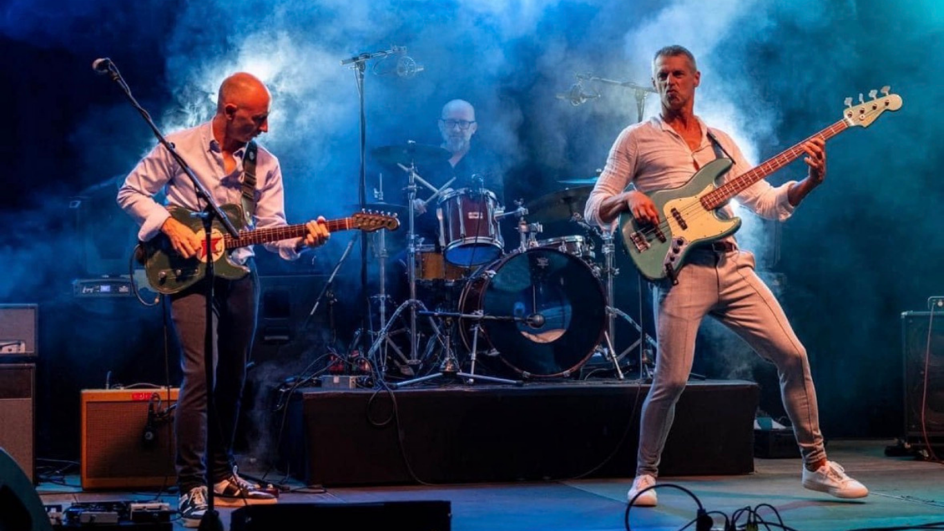 Three musicians performing on stage with colorful lighting and smoke, playing guitar, bass, and drums.