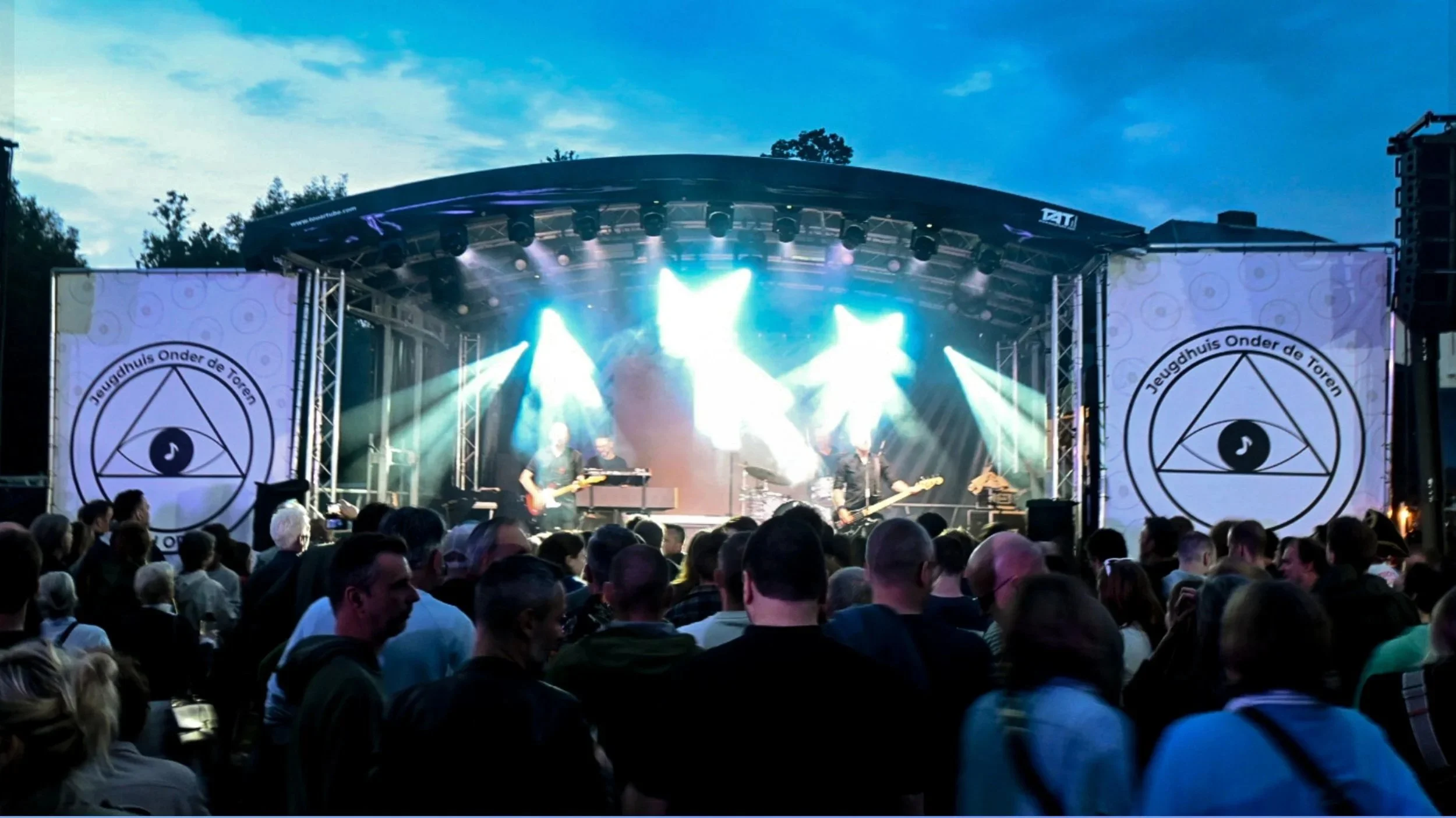 An outdoor concert stage with band performing under a evening sky, audience in foreground, stage decorated with banners displaying a geometric eye symbol and the text 'Jeugdhuis Onder de Toren'.