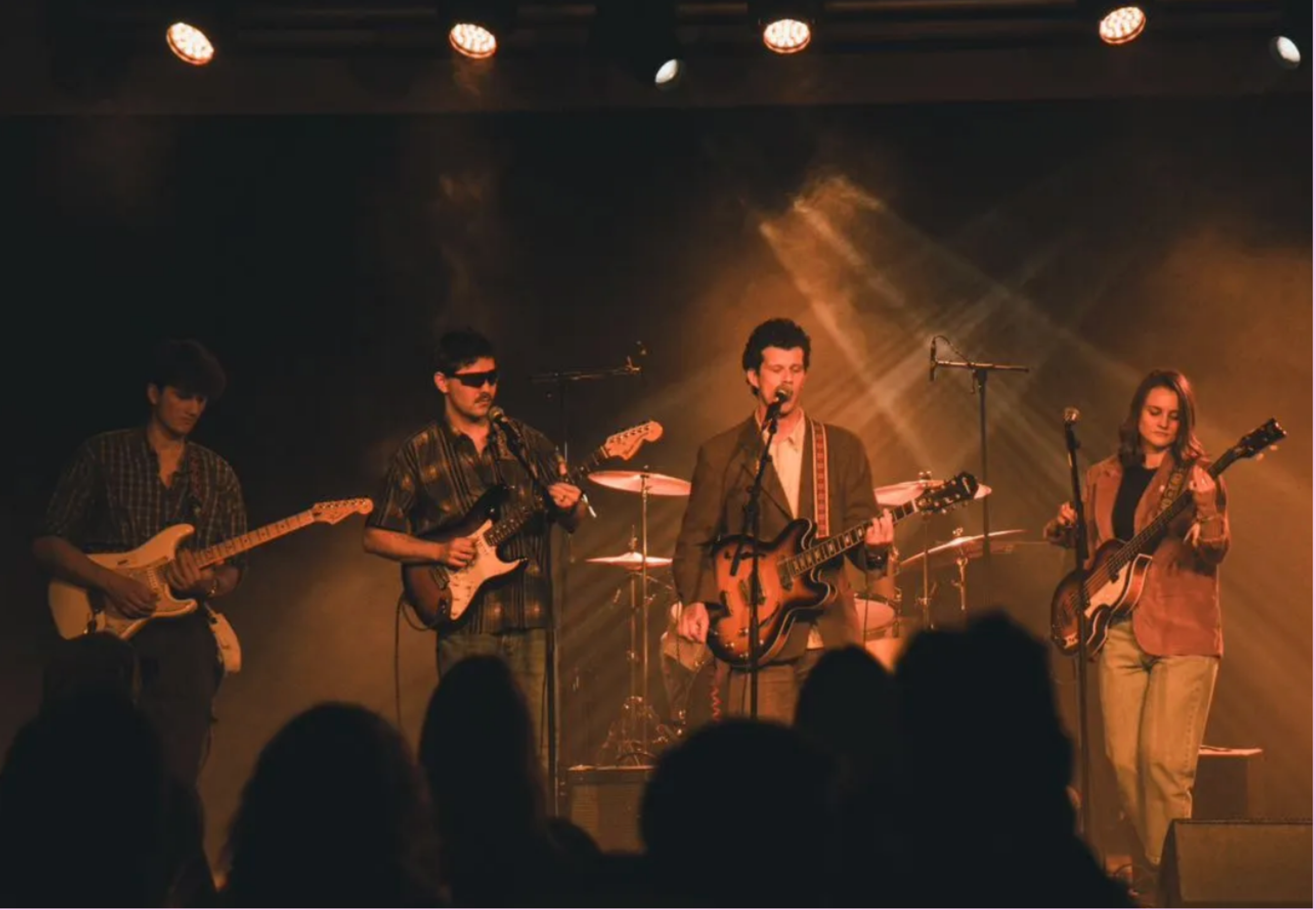 A band of five musicians performing on stage with guitars and drums under warm stage lighting, with an audience in front.