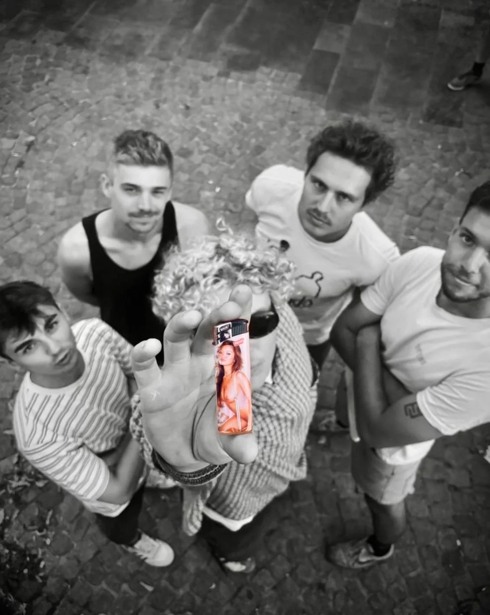 A group of five young men standing on a cobblestone street, with the central figure holding a lit lighter with a woman on it. The photo is black and white except for the lighter, which has a colorful image of a woman.