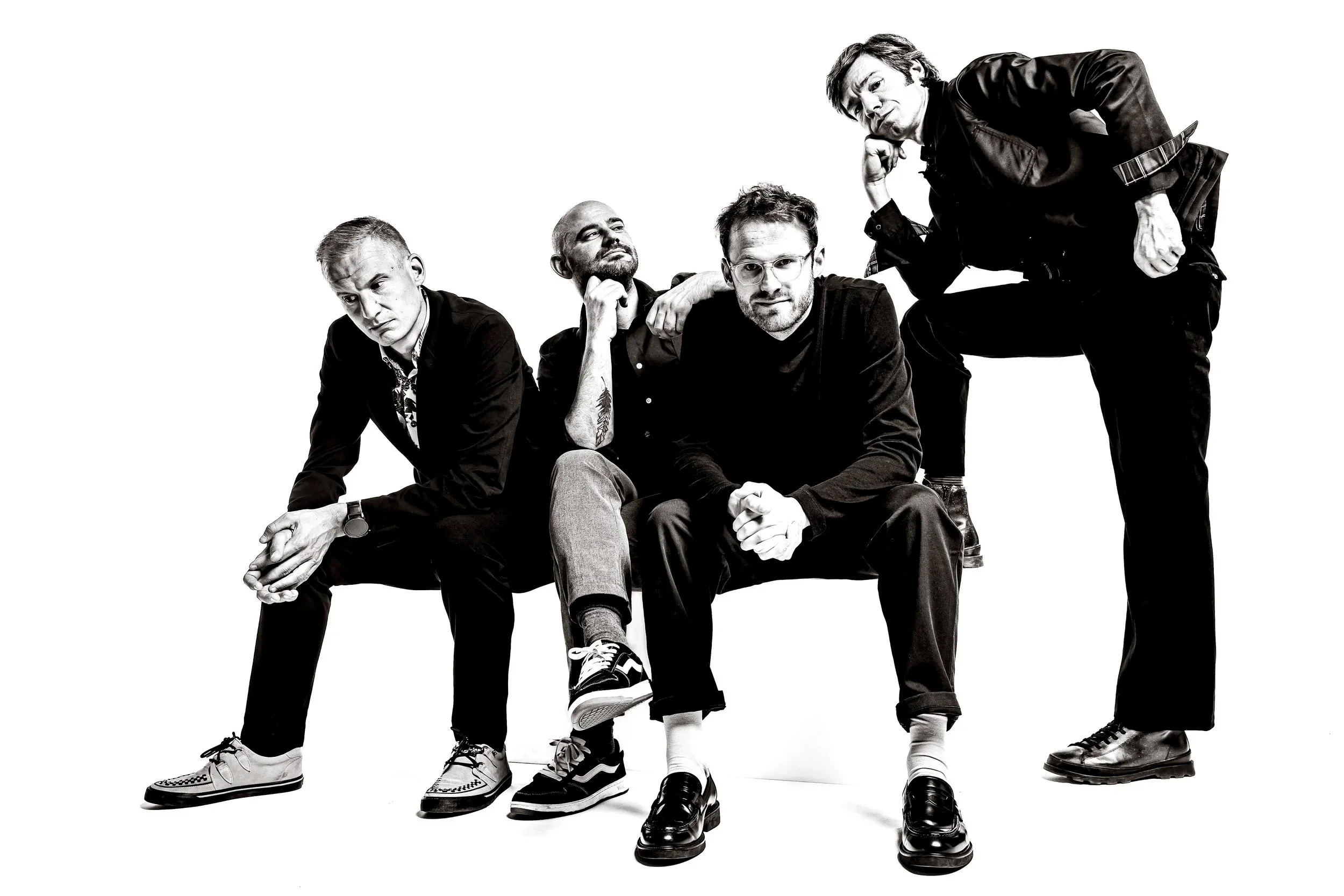 Black and white photo of five men posing casually, some sitting and some standing, with one man sitting on another man's knee, all looking at different angles.