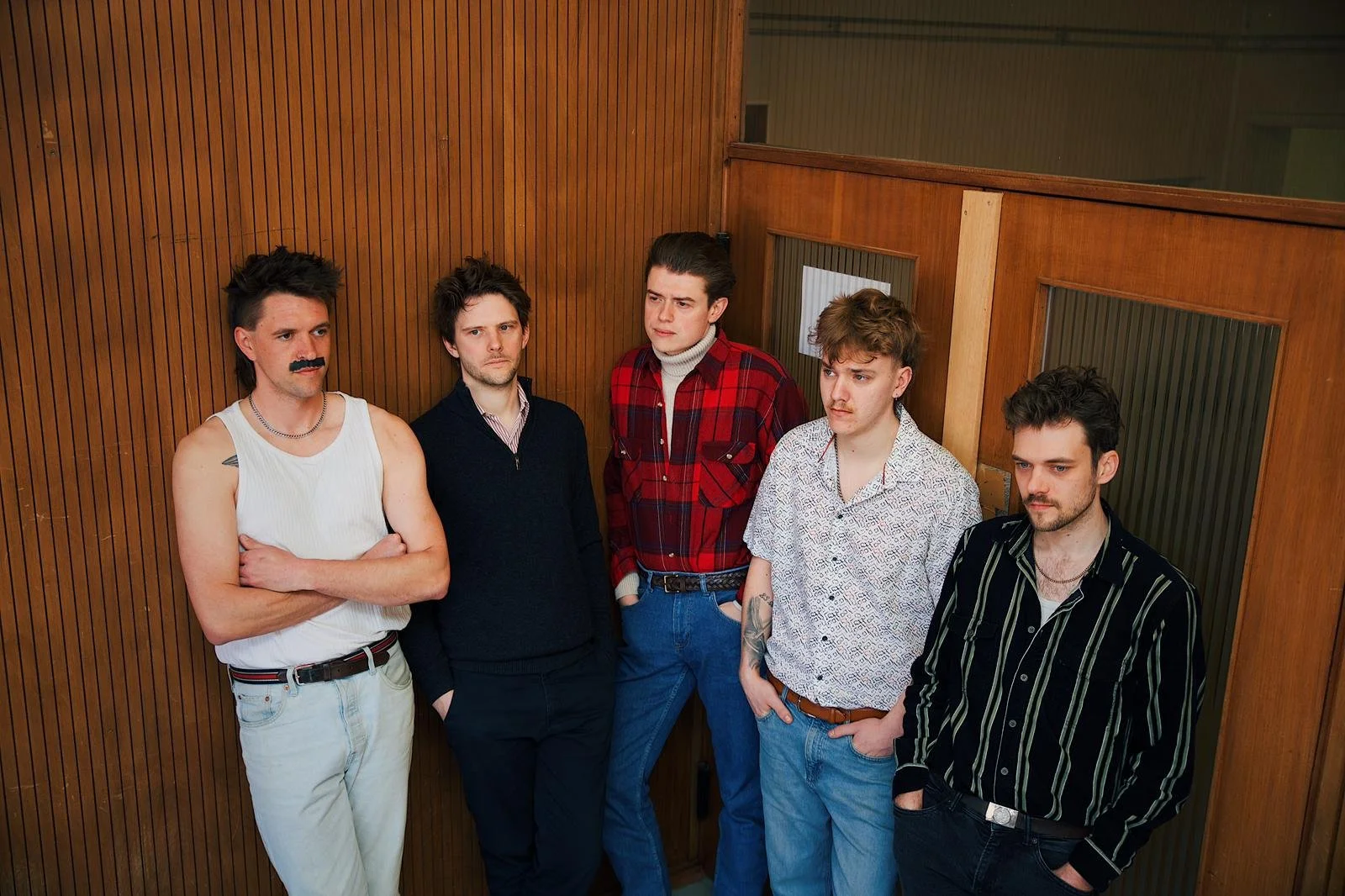 Five young men stand against wooden wall paneling in a narrow hallway, wearing casual 1980s-1990s style clothing, with serious expressions.