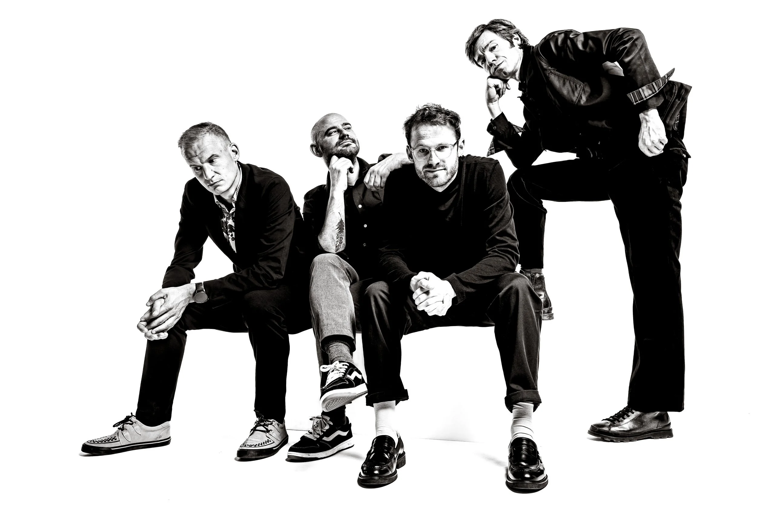Black-and-white photo of five men with diverse expressions and hairstyles posing together in a studio. Some are sitting or kneeling, while one is standing.
