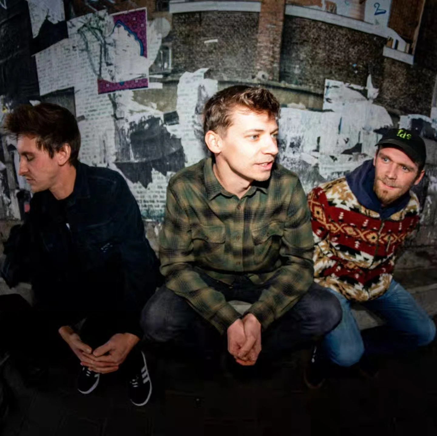 Three men seated in front of a torn wall covered with layered posters and graffiti, engaged in conversation, wearing casual clothing including a plaid shirt, a black jacket, and a patterned sweater.