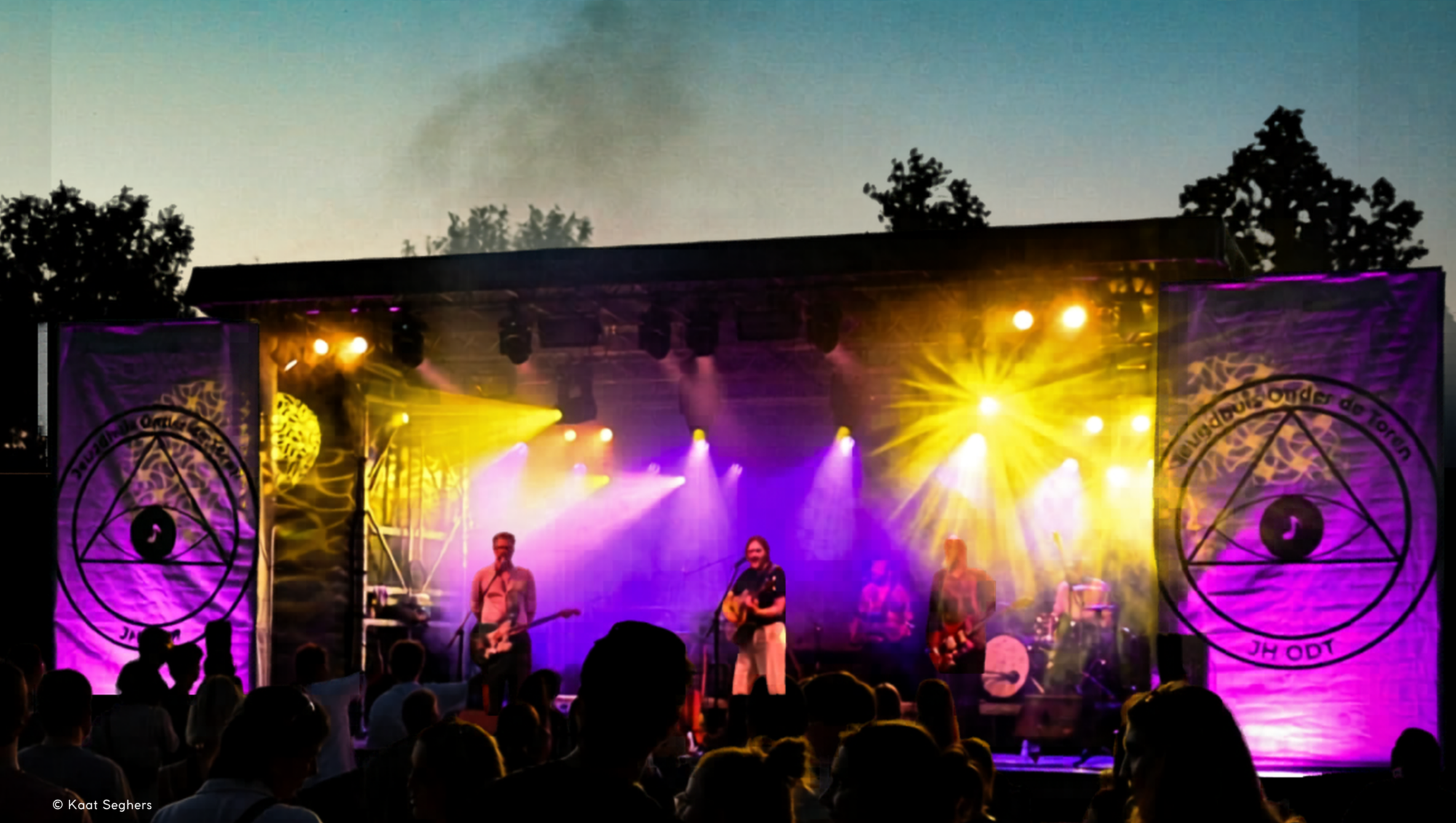 An outdoor concert at dusk with a stage illuminated by colorful yellow, purple, and pink lights. Several musicians are performing, and an audience is watching in the foreground. The backdrop features banners with circular designs and symbols, and trees are visible in the background against the evening sky.