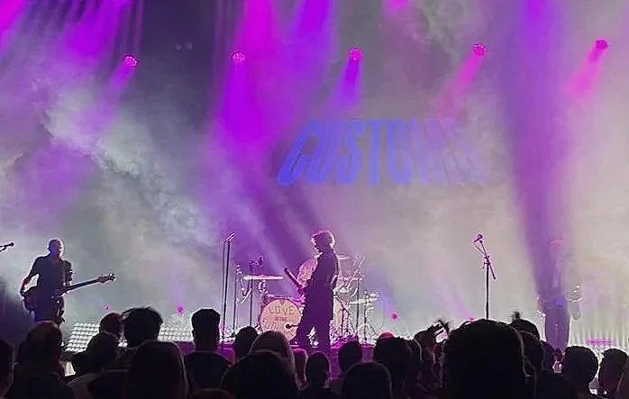 A band performing on stage with pink and purple lights, with a crowd watching. The background shows a large screen with the word "EASTTONE".