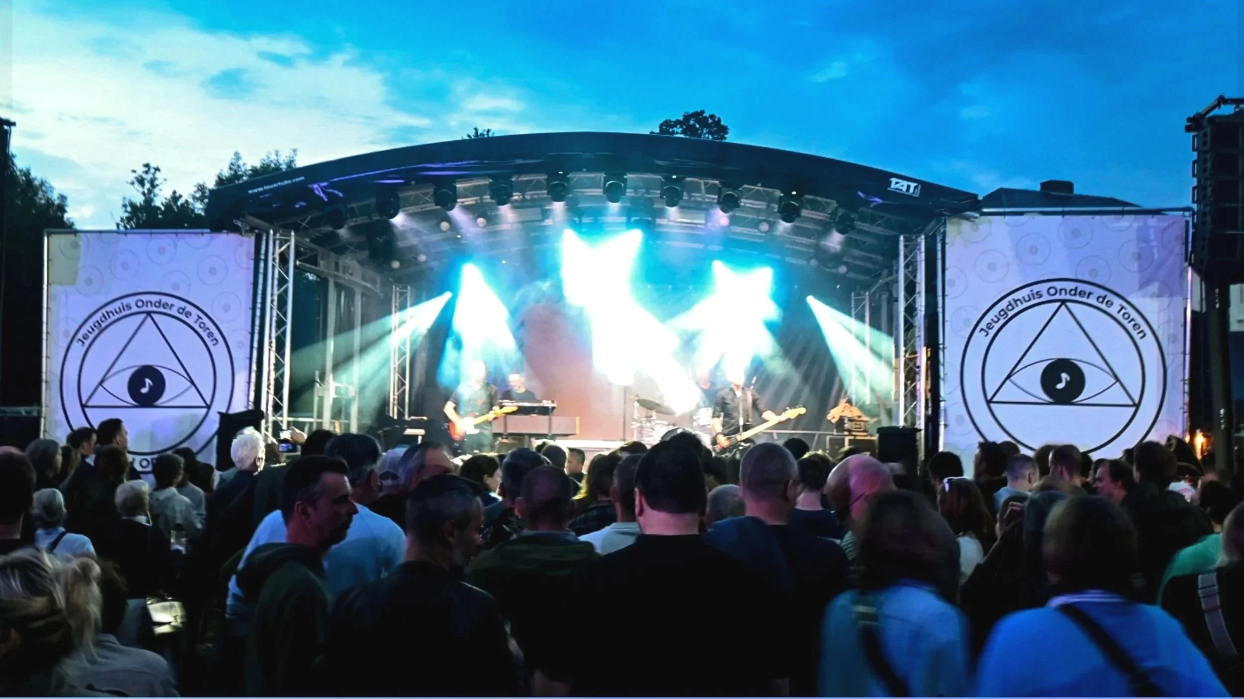 An outdoor concert with a large crowd watching a band perform on a stage illuminated with bright lights, with a blue sky and some clouds in the background. The stage has large screens with a black and white logo and the band members are playing guitars and drums.