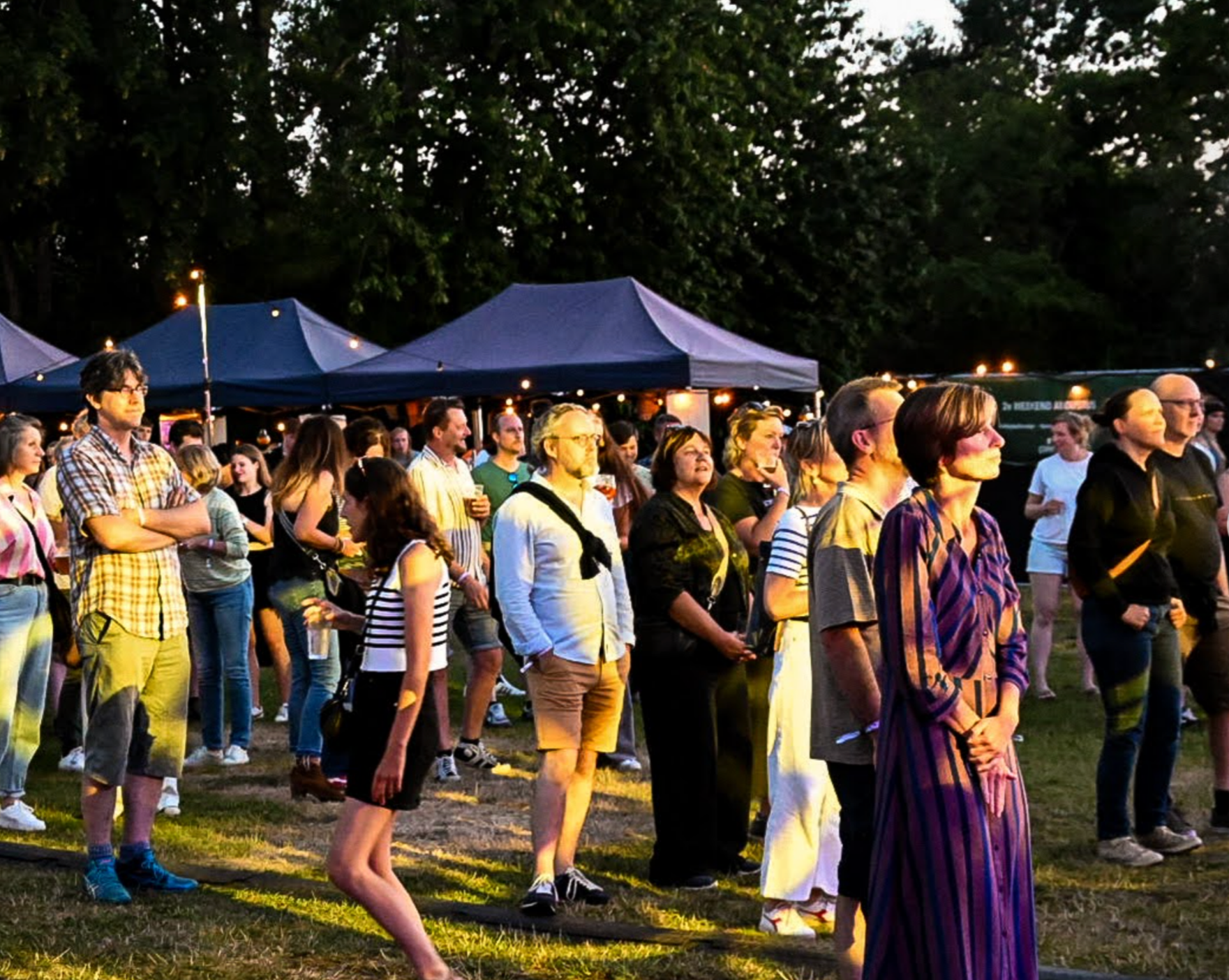 Gezicht op een menigte mensen die samen naar een podium kijken tijdens een avondevenement, met lichtjes en festivaltentagen op de achtergrond.