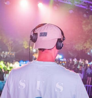 Back view of a person wearing a baseball cap and headphones at an outdoor concert or festival with colorful stage lights.