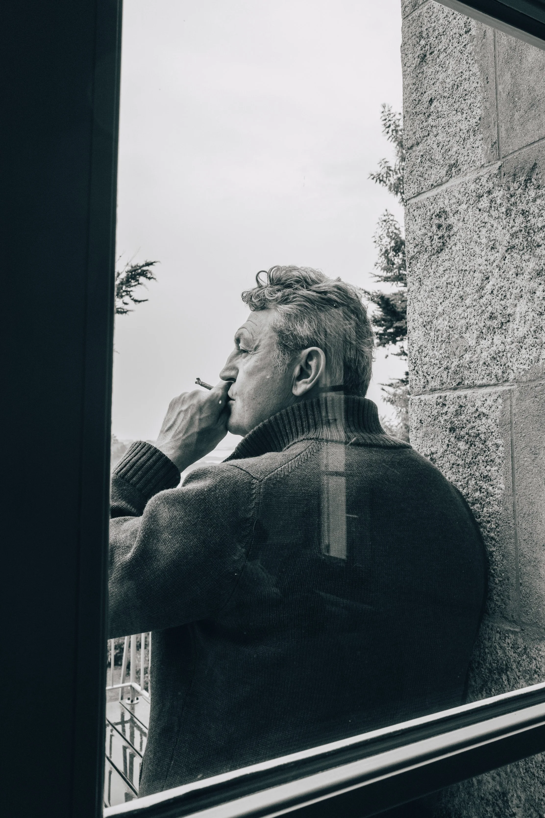 Un homme âgé, vu en profil, portant un pull à col roulé, fumant une cigarette, photographié par une fenêtre.