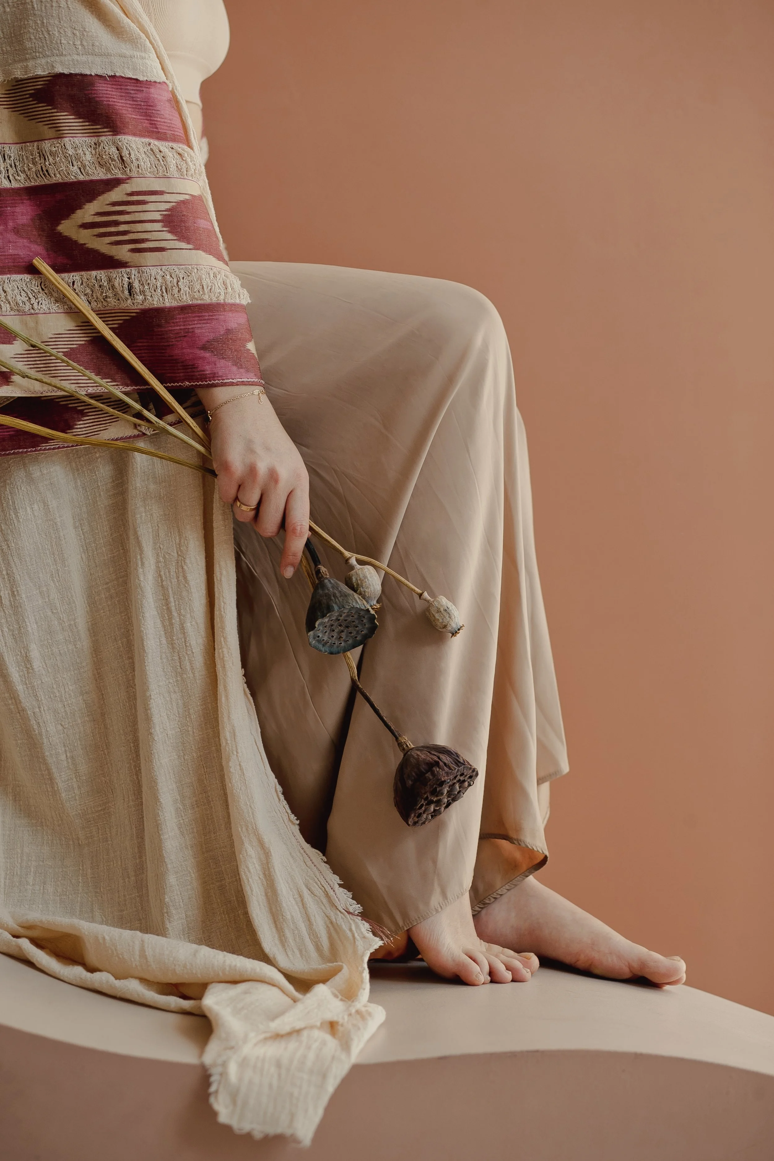 Woman in a flowing cream outfit seated barefoot, holding a dried flower