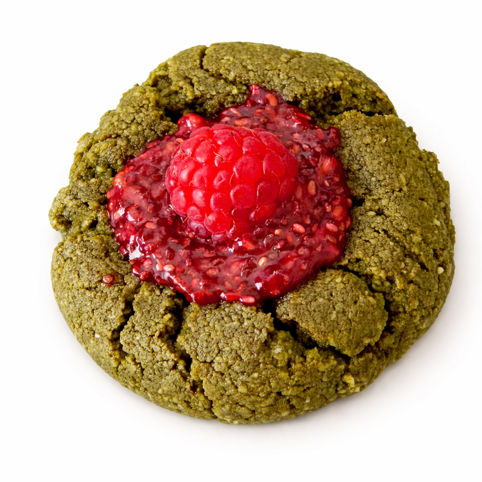 A green cookie topped with raspberry jam and a fresh raspberry on top, on a white background.