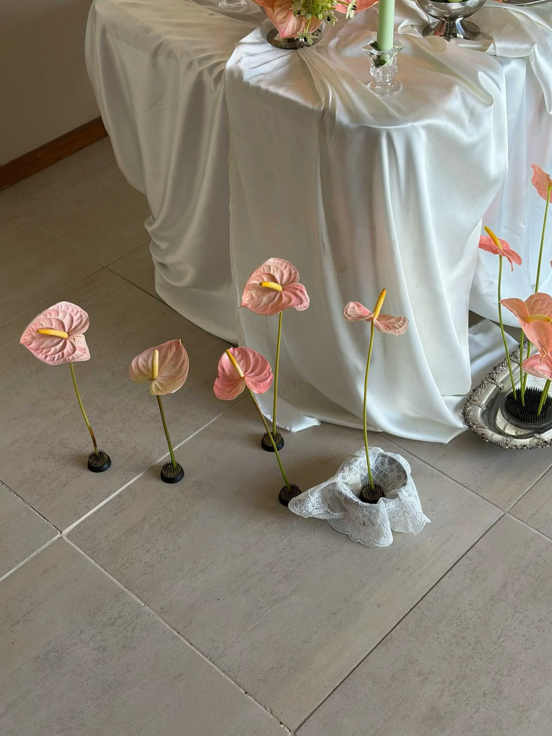 Arrangement of pink anthurium flowers with yellow spadices on a tiled floor, near a white-draped table with a silver tray holding additional flowers and candles.