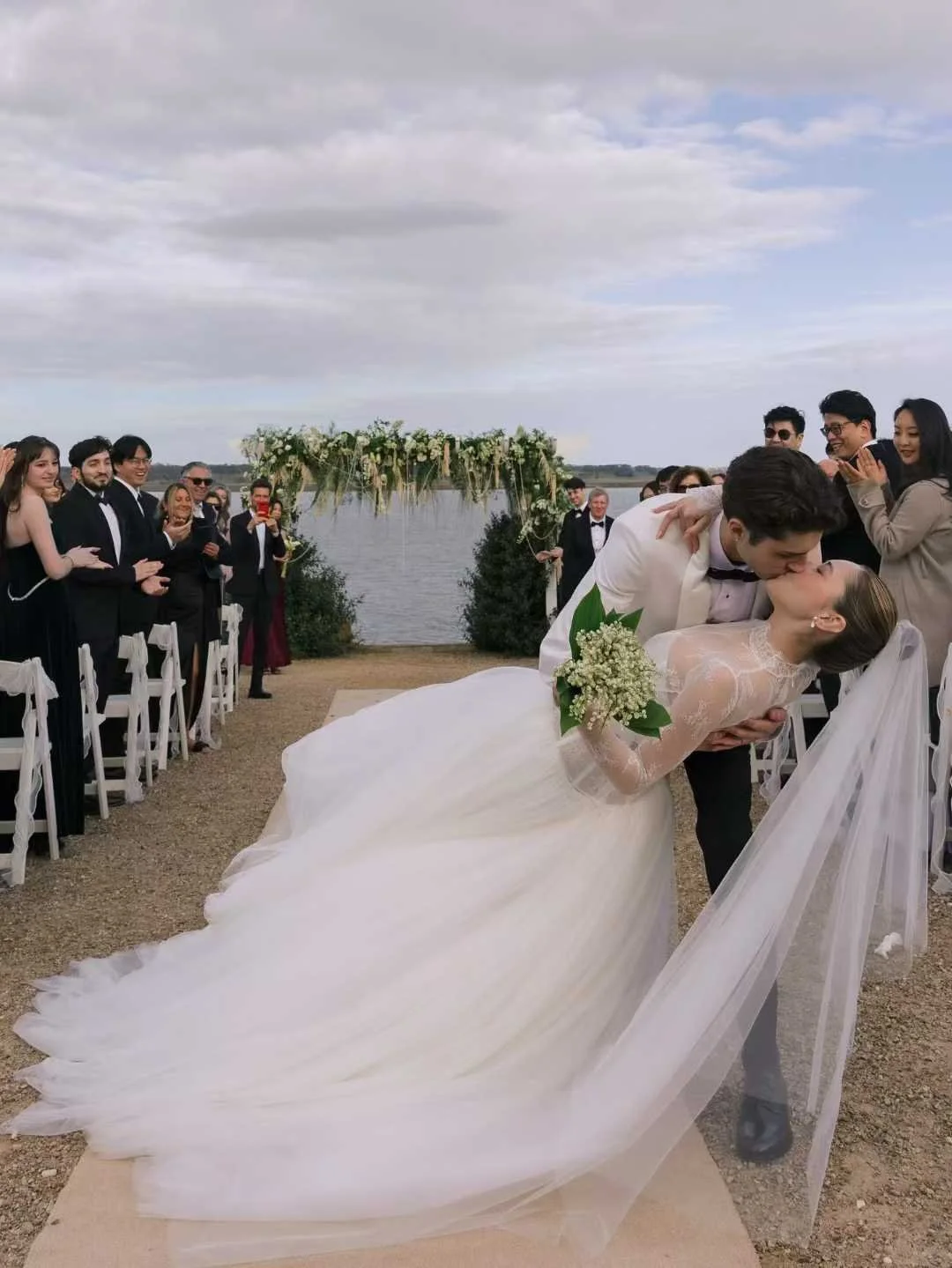 A newlywed couple shares a kiss during their wedding ceremony outdoors by a lake, with guests clapping and taking photos, and a floral arch in the background under a cloudy sky.