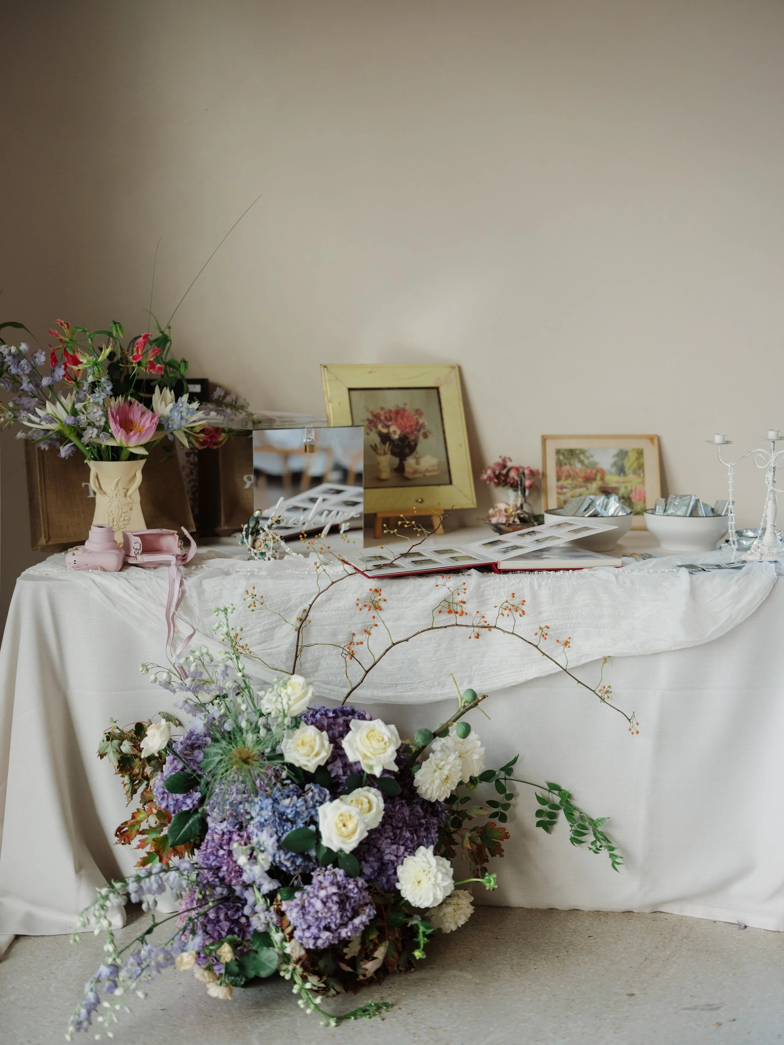 A table covered with a white cloth displaying framed art, bowls, and decorative items, with a floral arrangement in front featuring white roses, purple hydrangeas, and greenery.