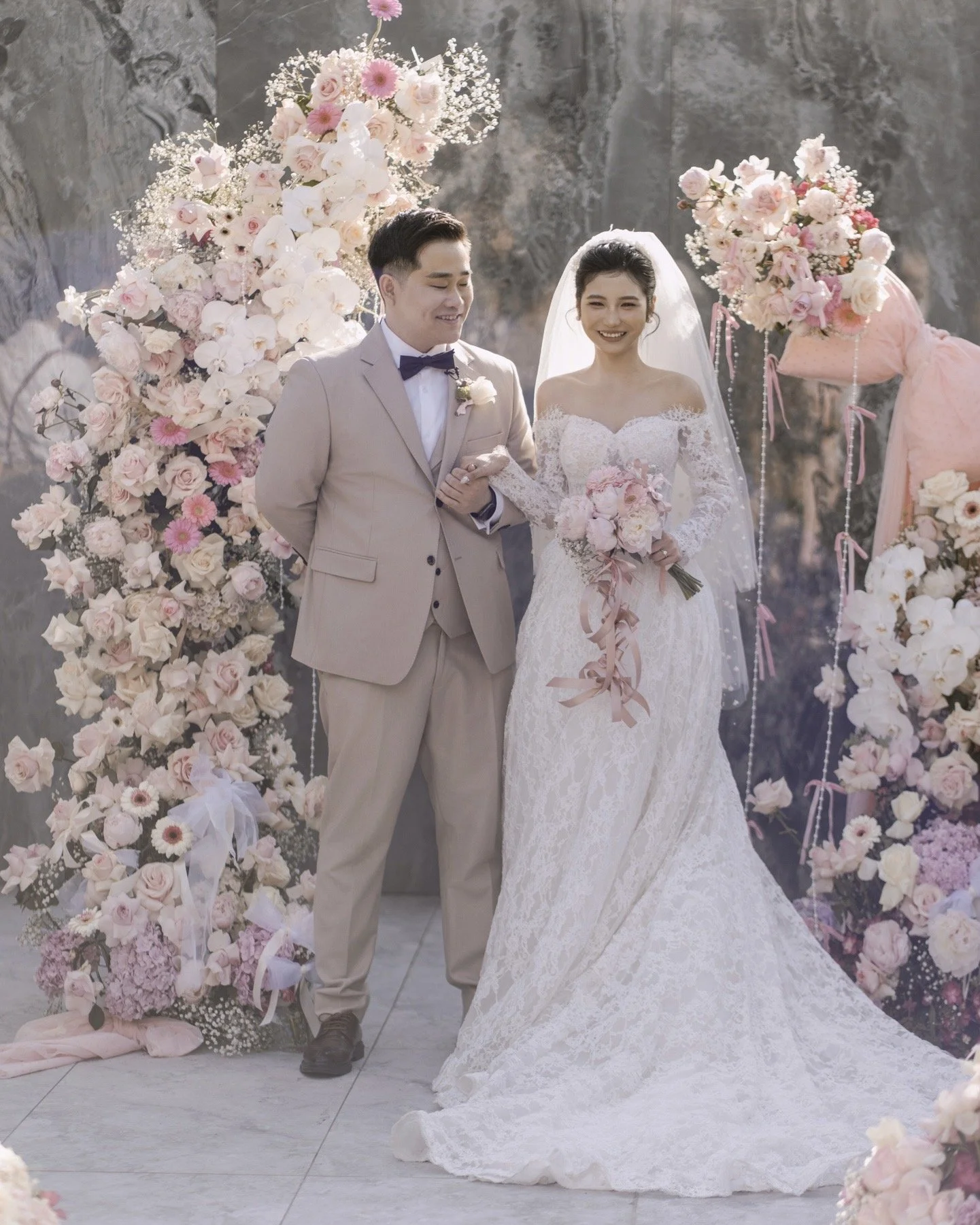 A bride and groom at their wedding ceremony, standing under floral arches with pink and white flowers.
