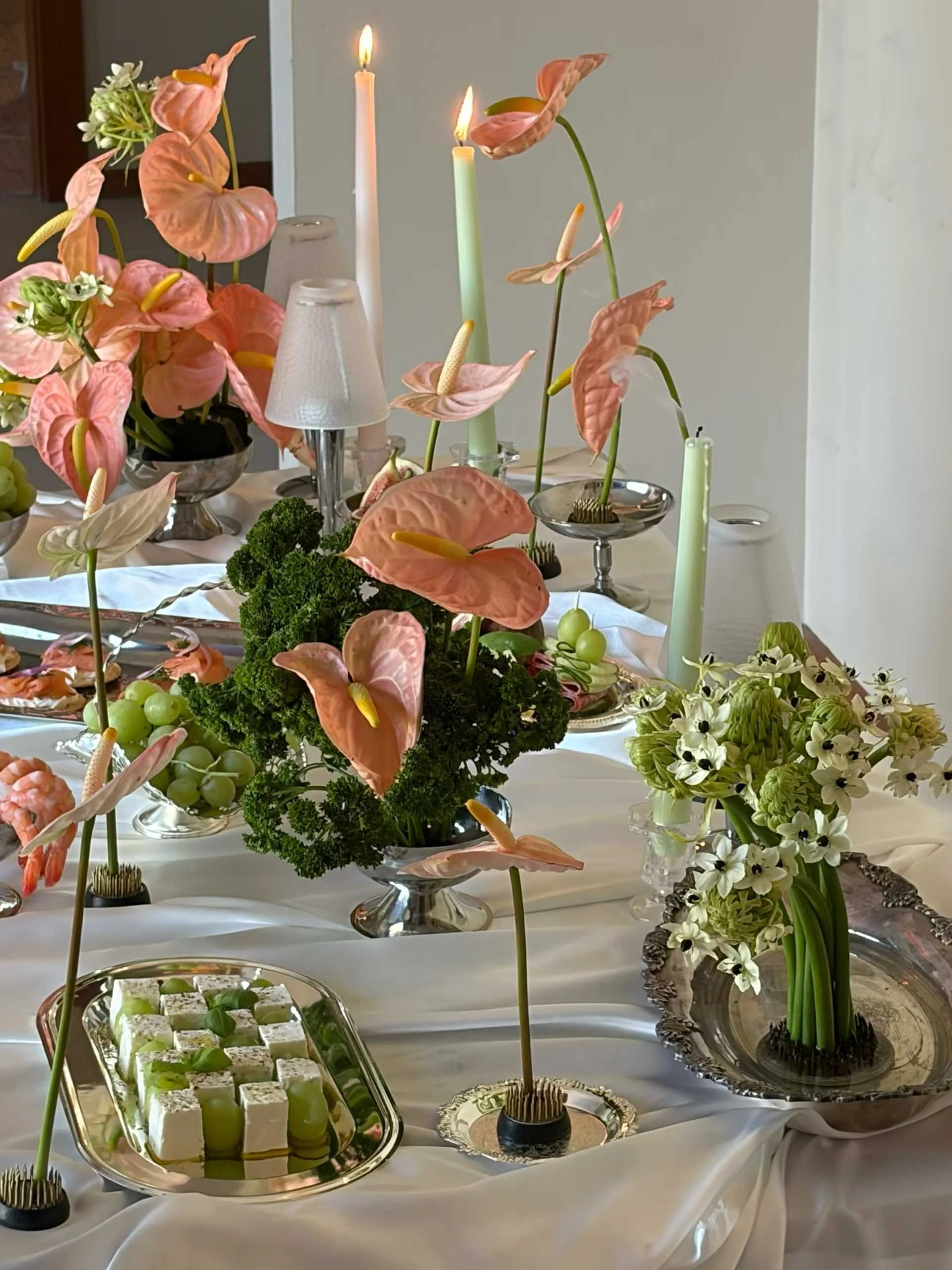 A table decorated with pink anthurium flowers, white candles, white and green floral arrangements, and a tray of green and white desserts, all on a white tablecloth.