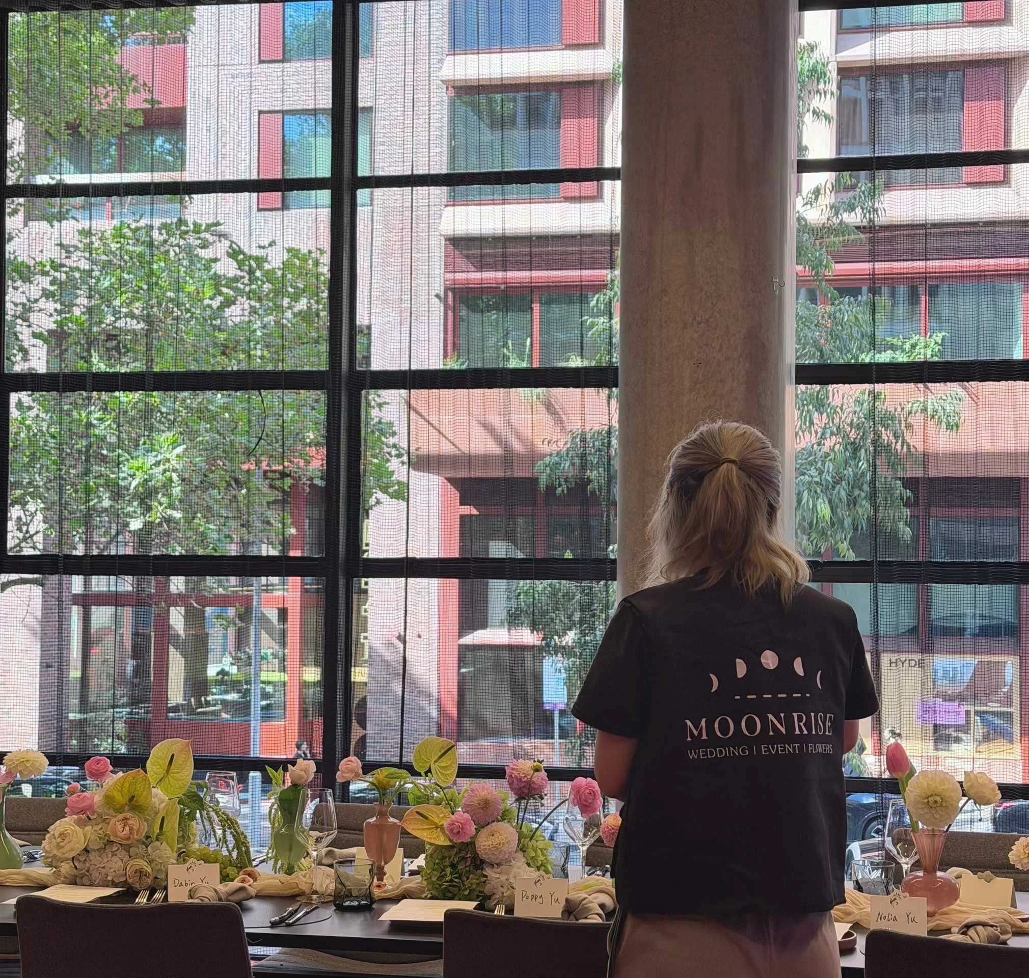 A woman standing at a decorated table with pink and white flowers inside a venue, looking out a large window at a city street with buildings and trees.