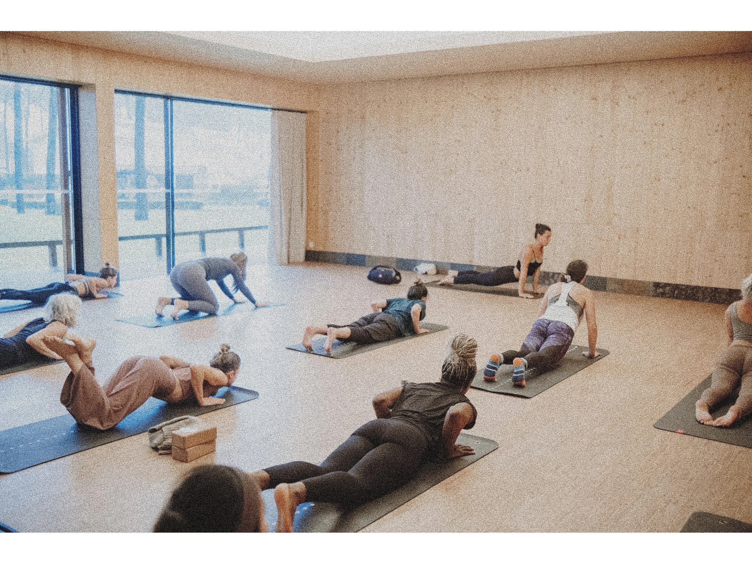 People participating in a group yoga class in a spacious, well-lit room with large windows.