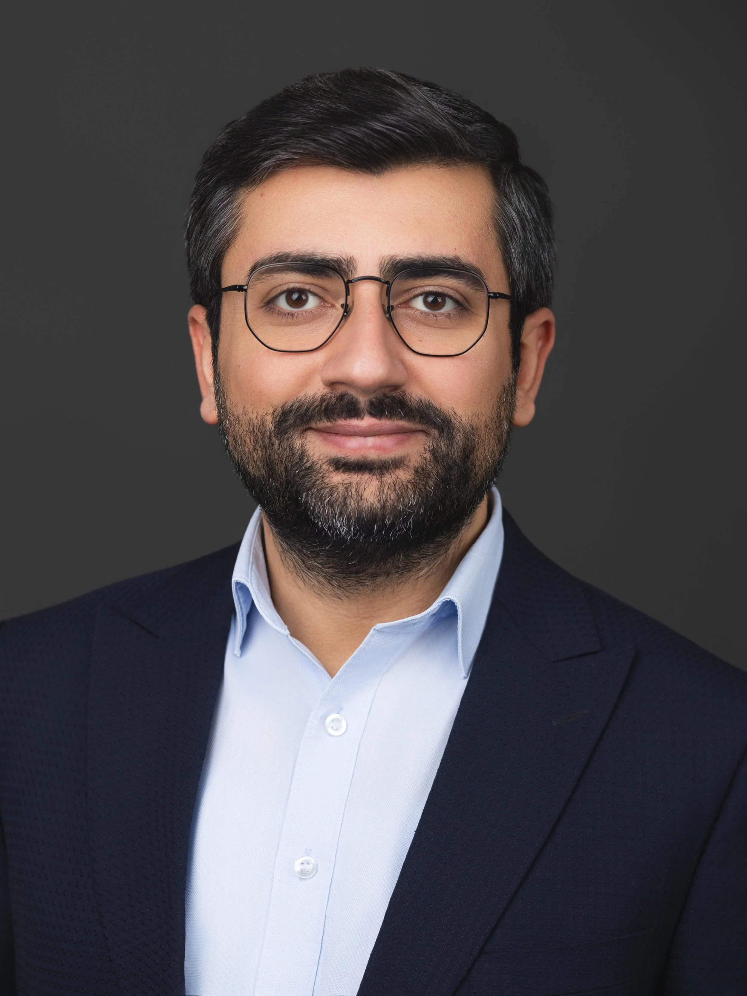 Headshot of a man with glasses, dark hair and beard, wearing a light blue shirt and a dark blazer, smiling against a dark gray background.