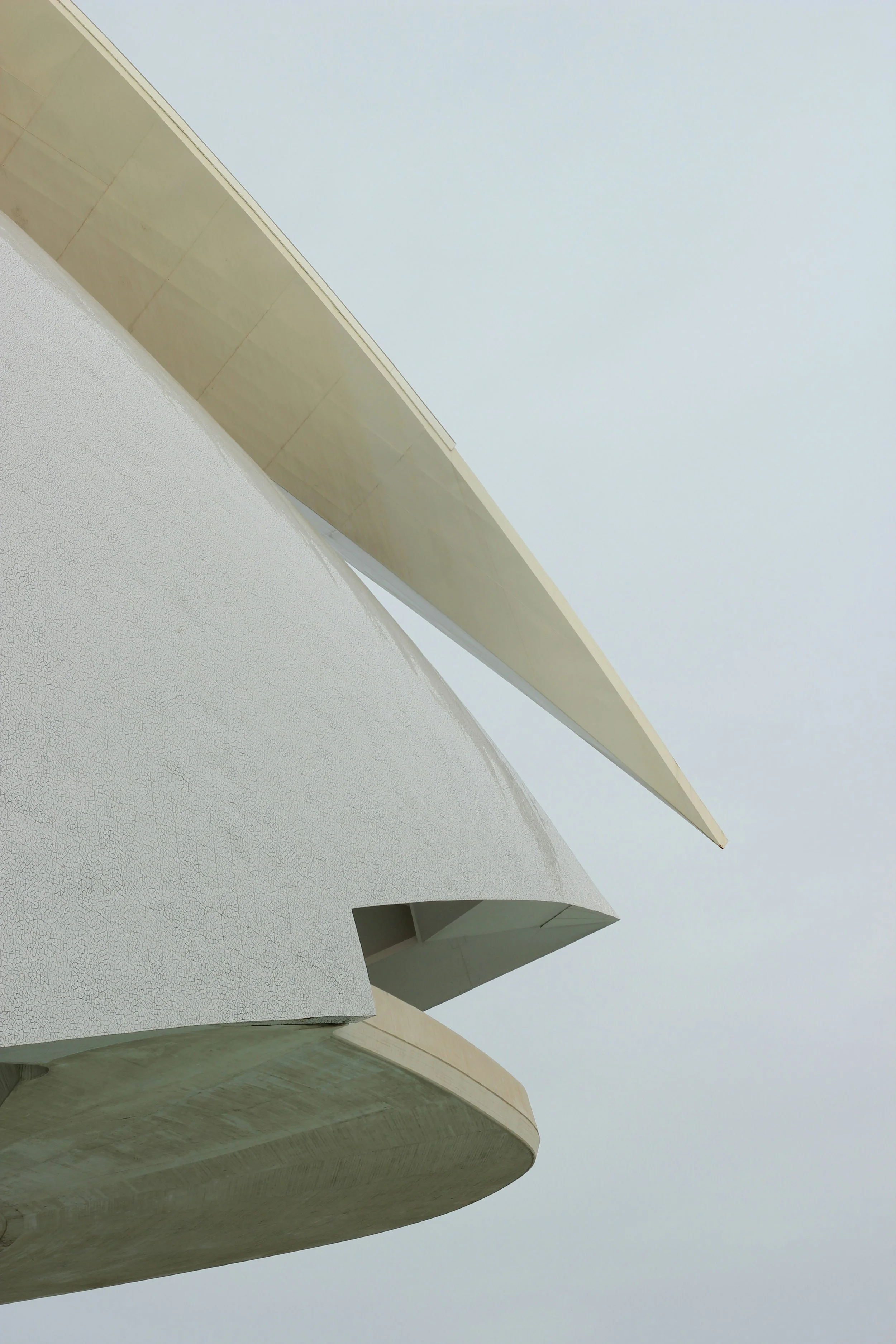 Close-up of the exterior of a modern building with curved white and beige concrete and a long beige overhang against a cloudy sky.