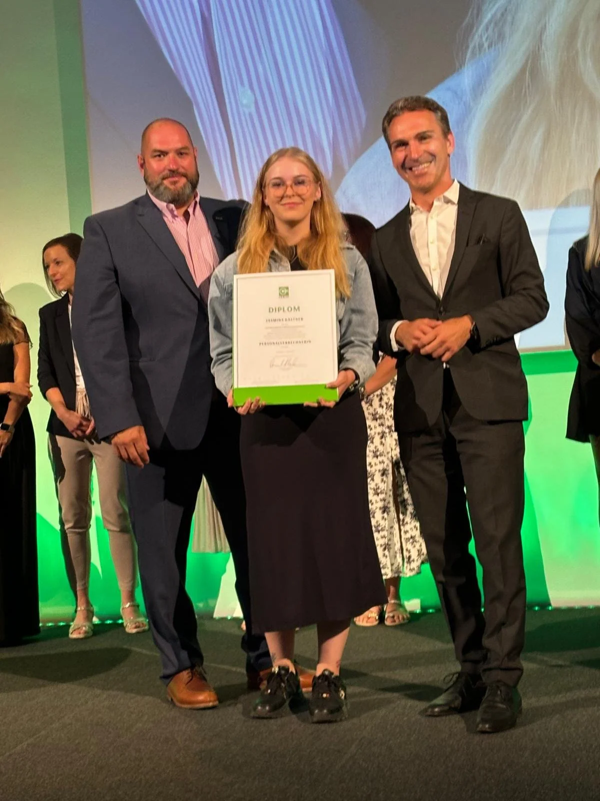 Eine Gruppe von Menschen bei einer Preisverleihung, zentral eine junge Frau mit Brille, die eine Urkunde hält, flankiert von zwei Männern in Anzügen.