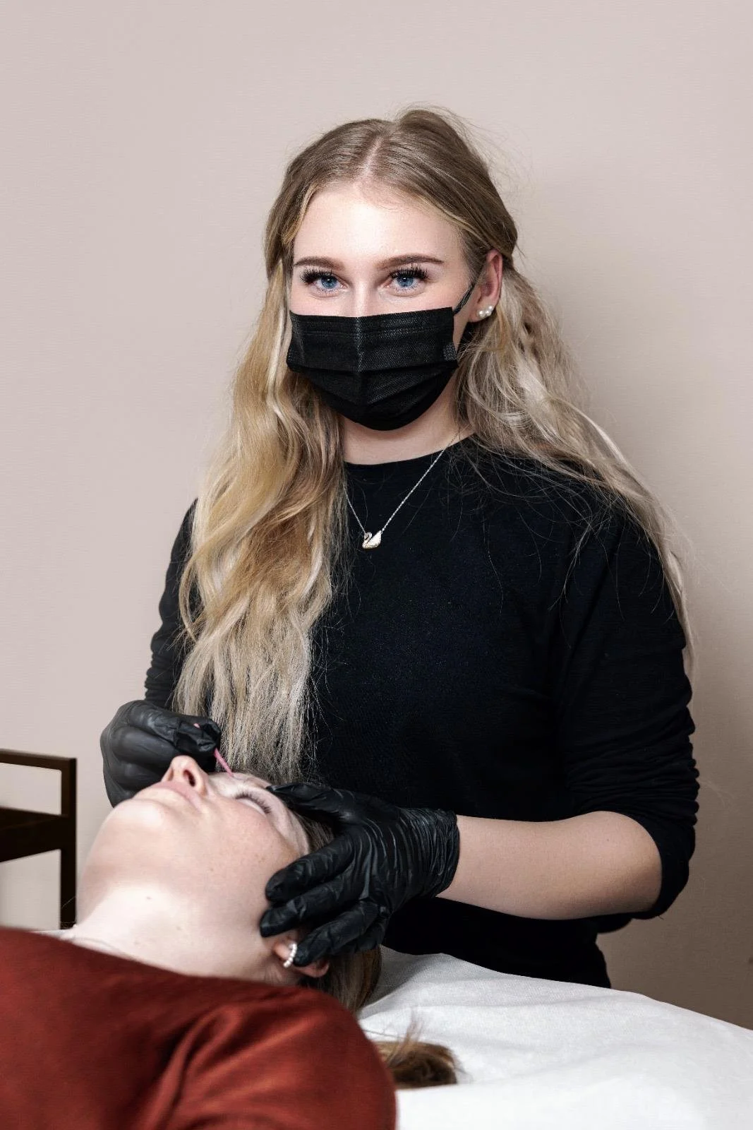 Eine Frau mit schwarzen Handschuhen führt eine kosmetische Behandlung im Gesicht einer anderen Frau durch, beide tragen schwarze Masken.