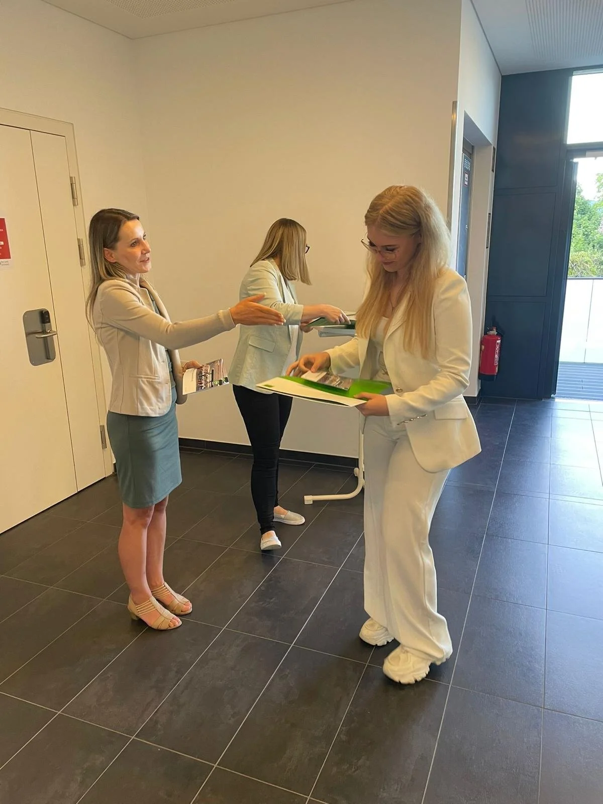 Drei Frauen stehen in einem Flur, eine hält einen Ordner, die anderen zwei geben sich Dokumente oder Informationsmaterial. Die Frauen sind formell gekleidet, eine ist in Weiß, die andere trägt hellgrau. Es scheint eine geschäftliche oder berufliche Situation zu sein.