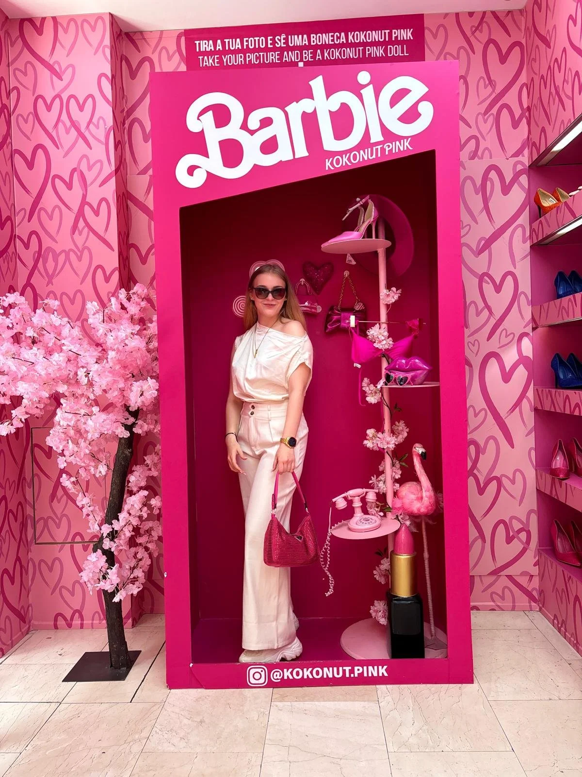 Eine Frau posiert vor einer pinken Barbie-Dekoration im CocoNUT PINK Laden, umgeben von pinken Herzen, Blumen und Barbie-Artikeln. Sie trägt eine weiße Kleidung, dunkle Sonnenbrille und hält eine pinke Tasche.