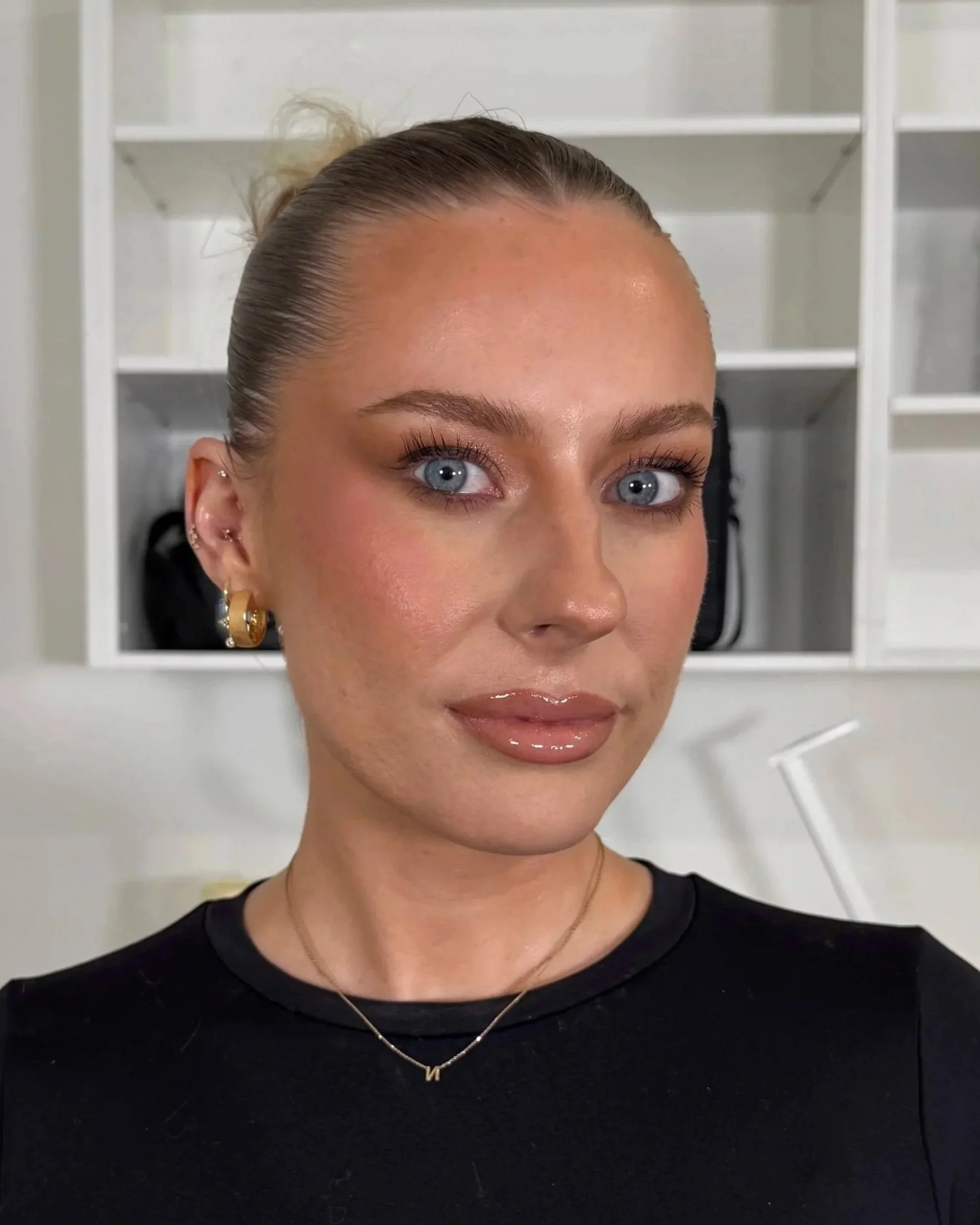 A woman with blonde hair wrapped in a bun, wearing gold hoop earrings, a black top, and a gold necklace with the letter 'M', standing indoors against white shelves.
