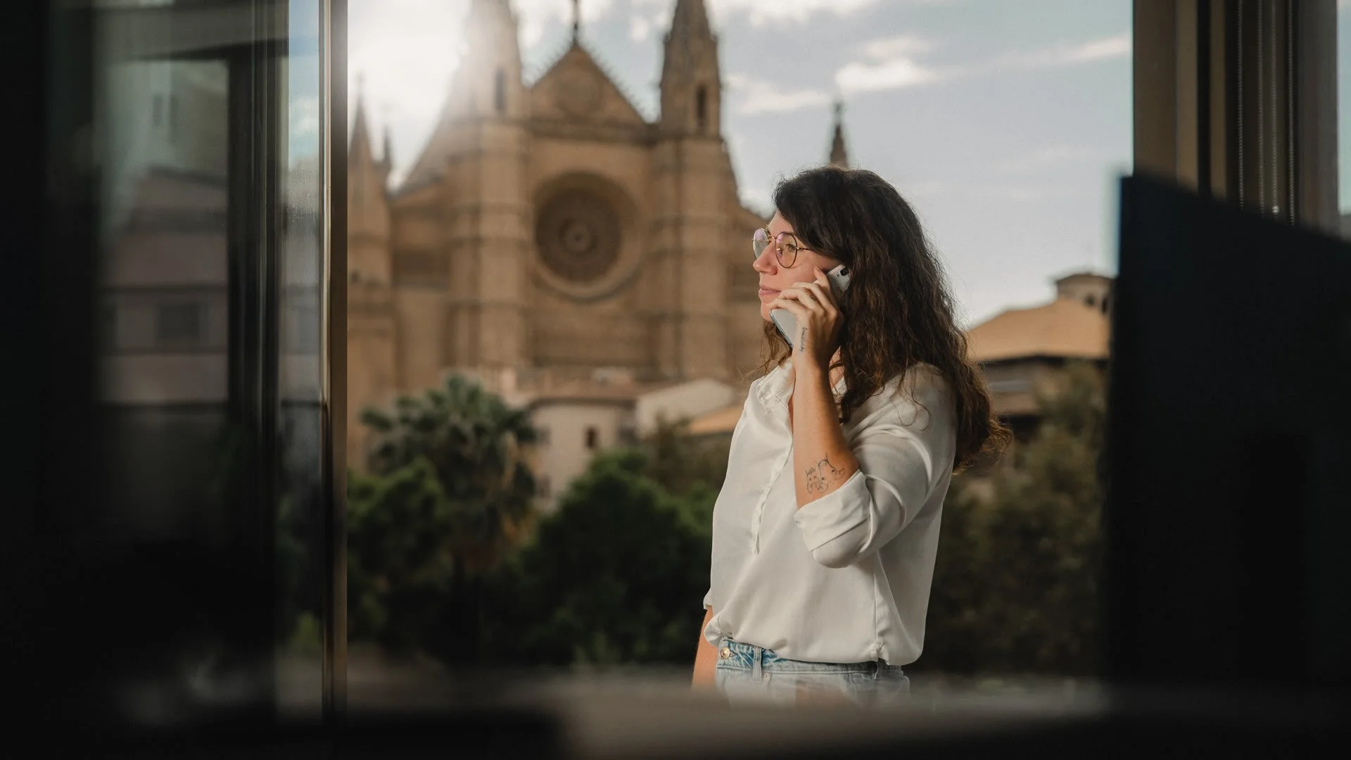 Mujer con gafas y tatuaje hablando por teléfono en un balcón, con la catedral de Palma visible en el fondo y un cielo parcialmente nublado.