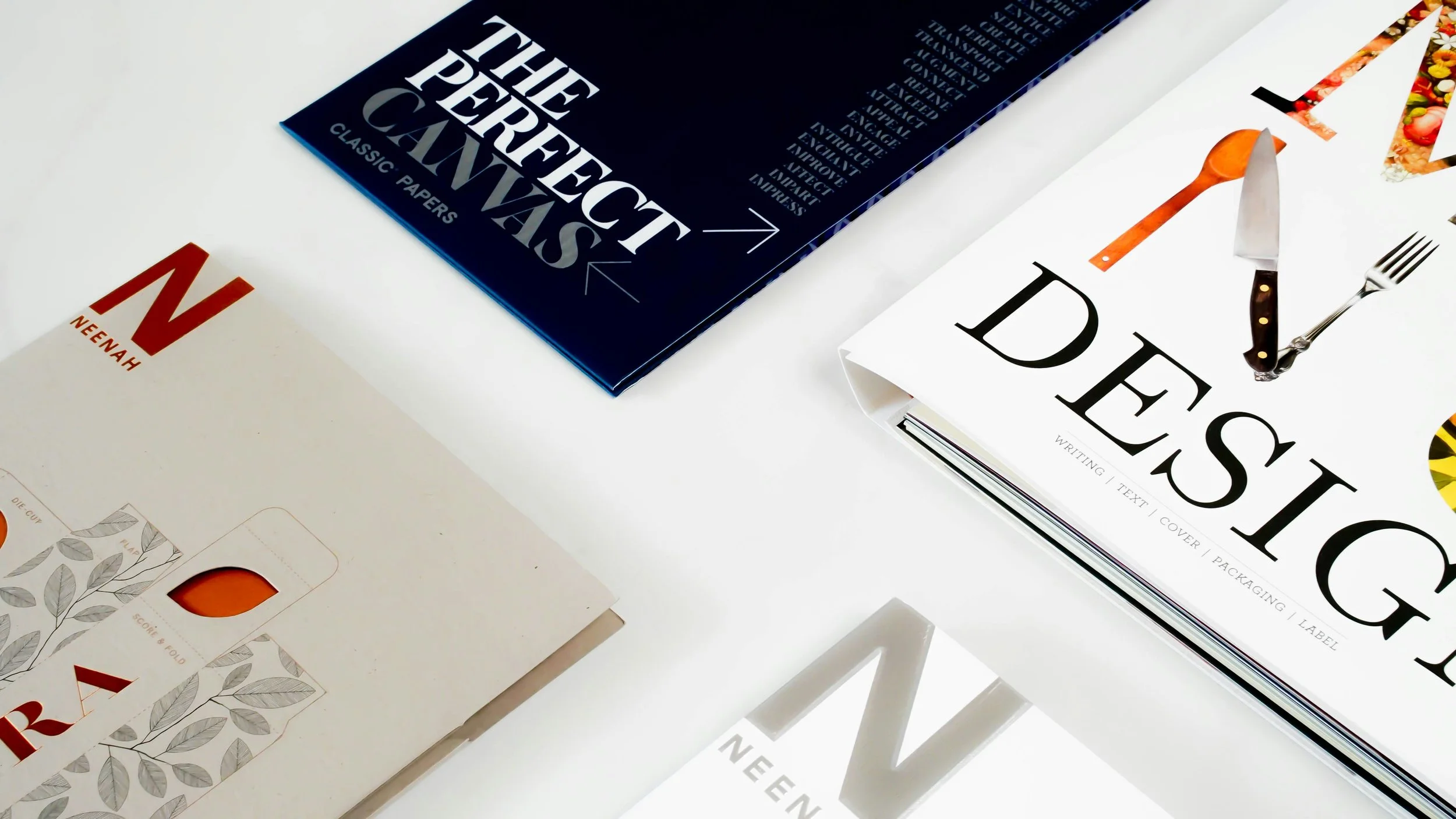 Libros y papeles en una mesa, incluyendo un libro azul oscuro titulado 'The Perfect Canvas', un libro blanco con letras grandes y una cuchara, un cuchillo y un tenedor sobre él, y tarjetas de papel con diseños botánicos.