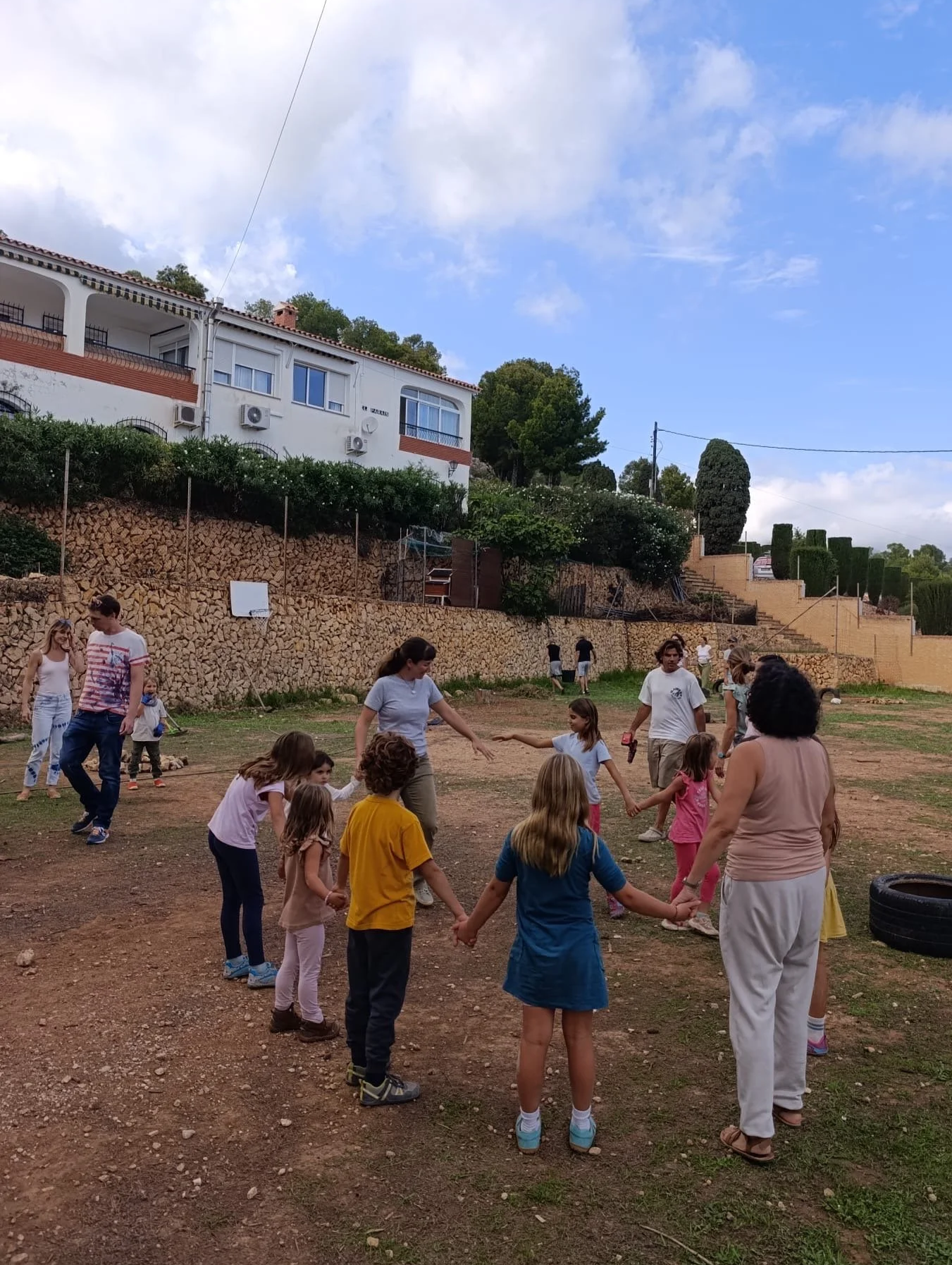 Grupo de niños y adultos jugando en un campo de tierra al aire libre, con casas y árboles en el fondo, en un día soleado y con algunas nubes.