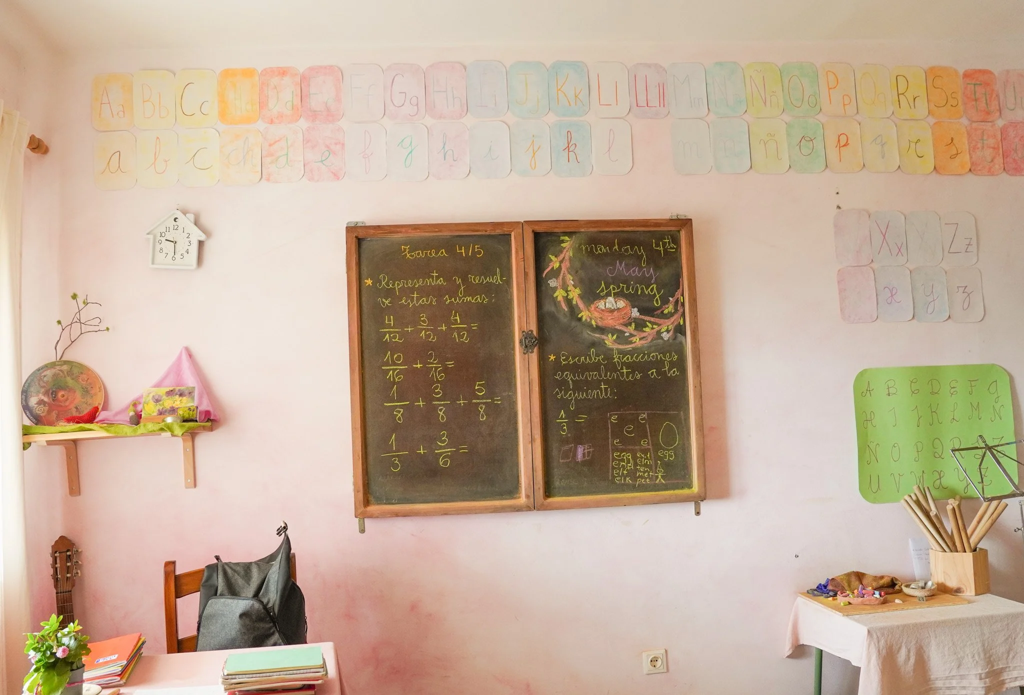 Aula escolar con paredes rosas, cartel con el alfabeto en diferentes colores, dos pizarras con ejercicios de fracciones y matemáticas, un reloj en forma de casa en la pared, mesas con libros y objetos escolares, y decoraciones variadas.