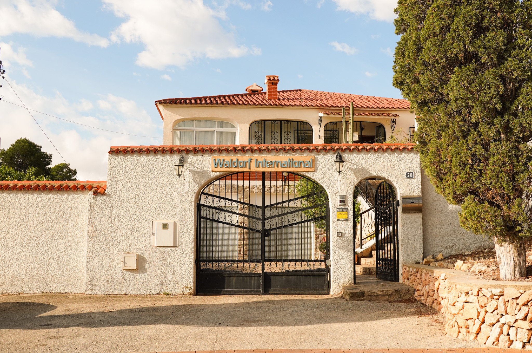 Entrada de una residencia con un muro blanco, portón de metal negro, un cartel que dice Waldorf International y un edificio de dos pisos con techo de tejas rojas y chimenea, rodeado de árboles verdes.