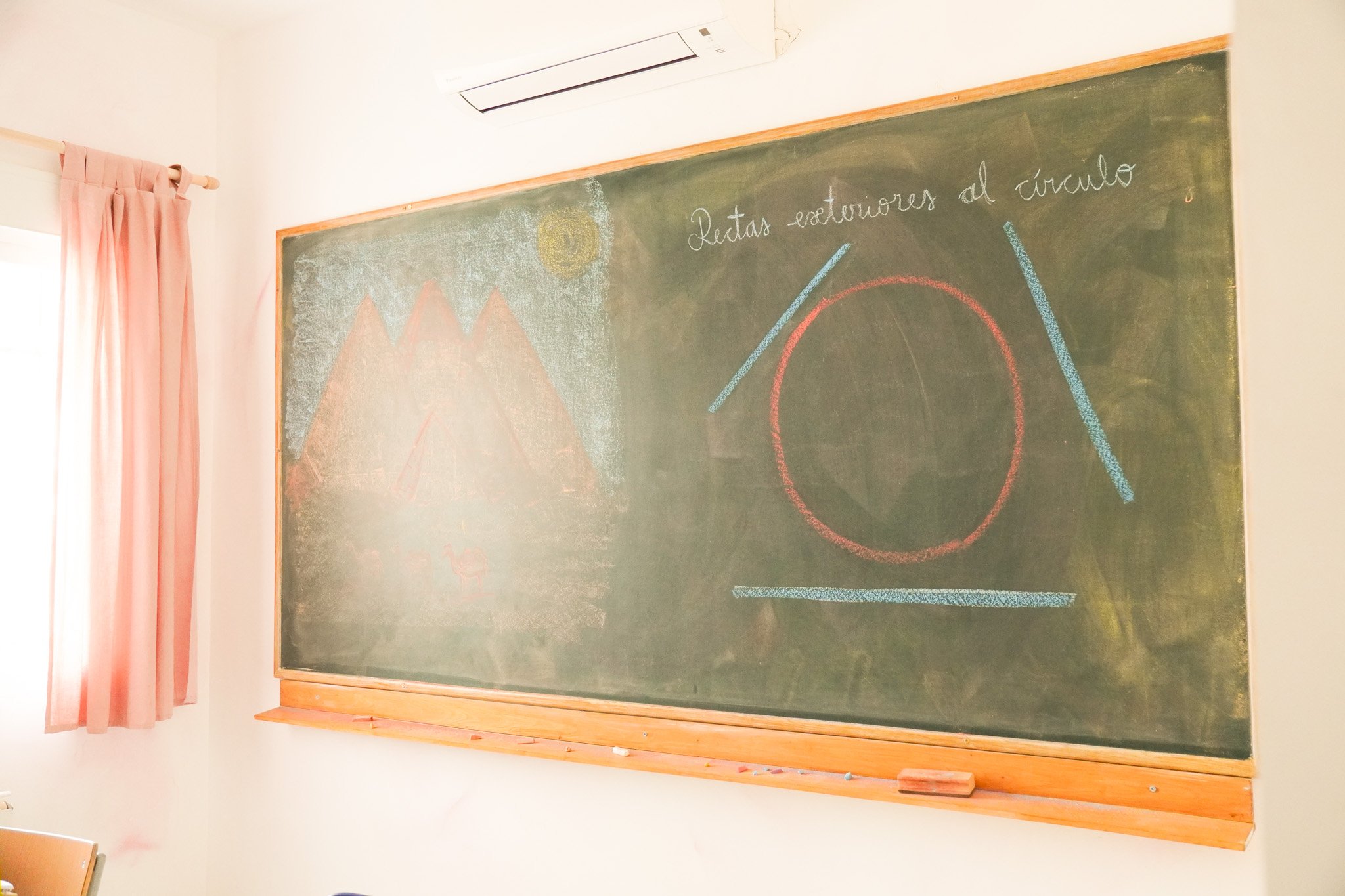 Pizarra de pizarra verde con instrucciones escritas en tiza blanca y algunos dibujos y marcas en diferentes colores, en una sala de clases con cortinas rosadas a la izquierda.