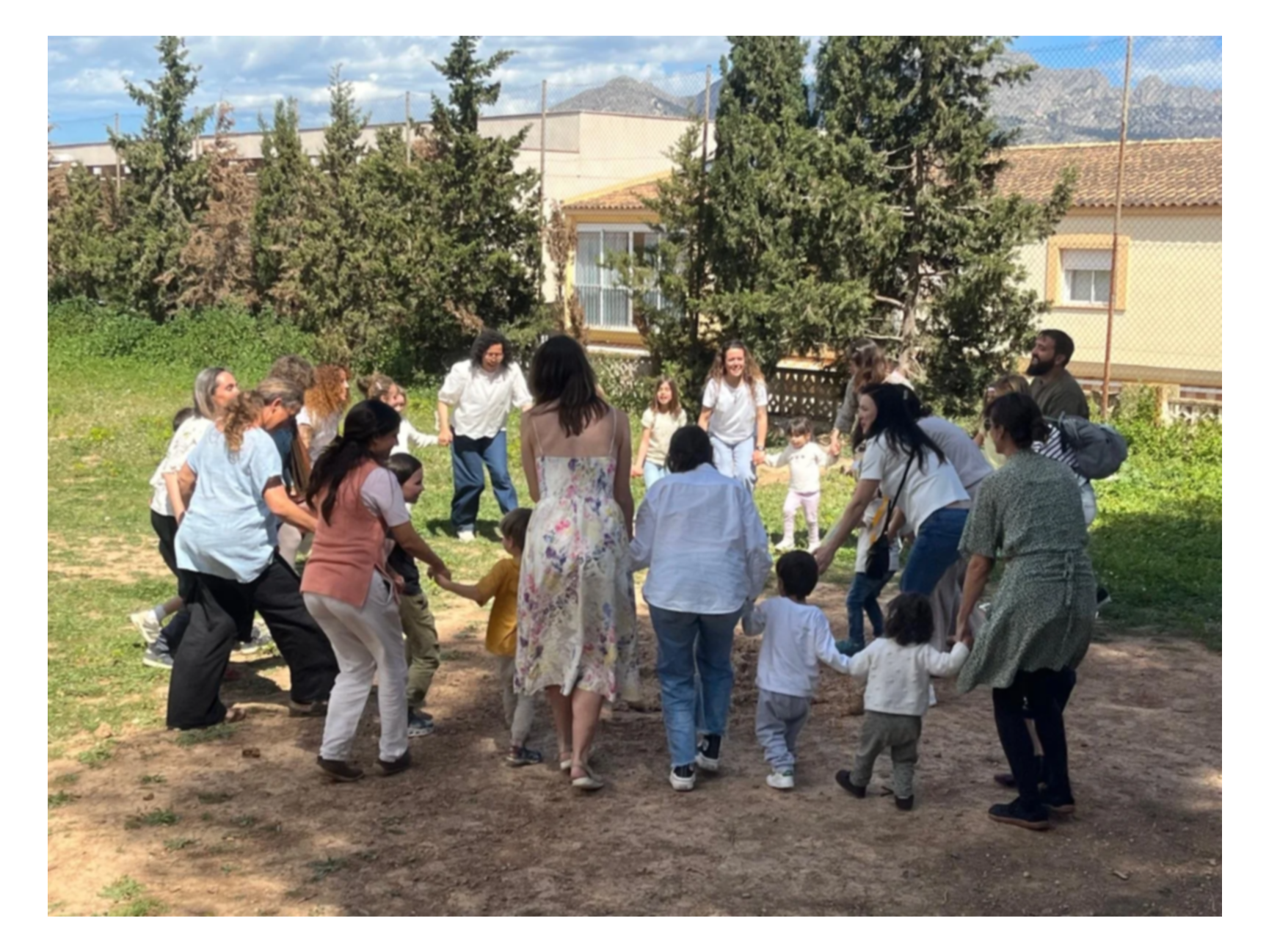 Grupo de personas, adultos y niños, formando un círculo y tomados de las manos en un espacio al aire libre con árboles, casas y montañas de fondo.