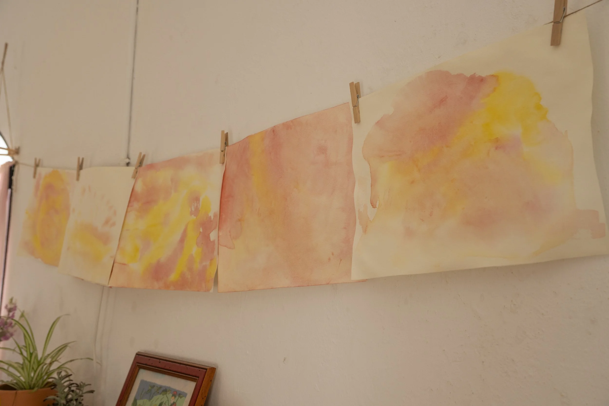 Pinturas abstractas de acuarela en tonos naranjas, rosas y amarillos colgadas en una cuerda con pinzas, en una pared blanca. En la parte inferior izquierda, una planta en una maceta y en la esquina inferior derecha, un cuadro colgado.