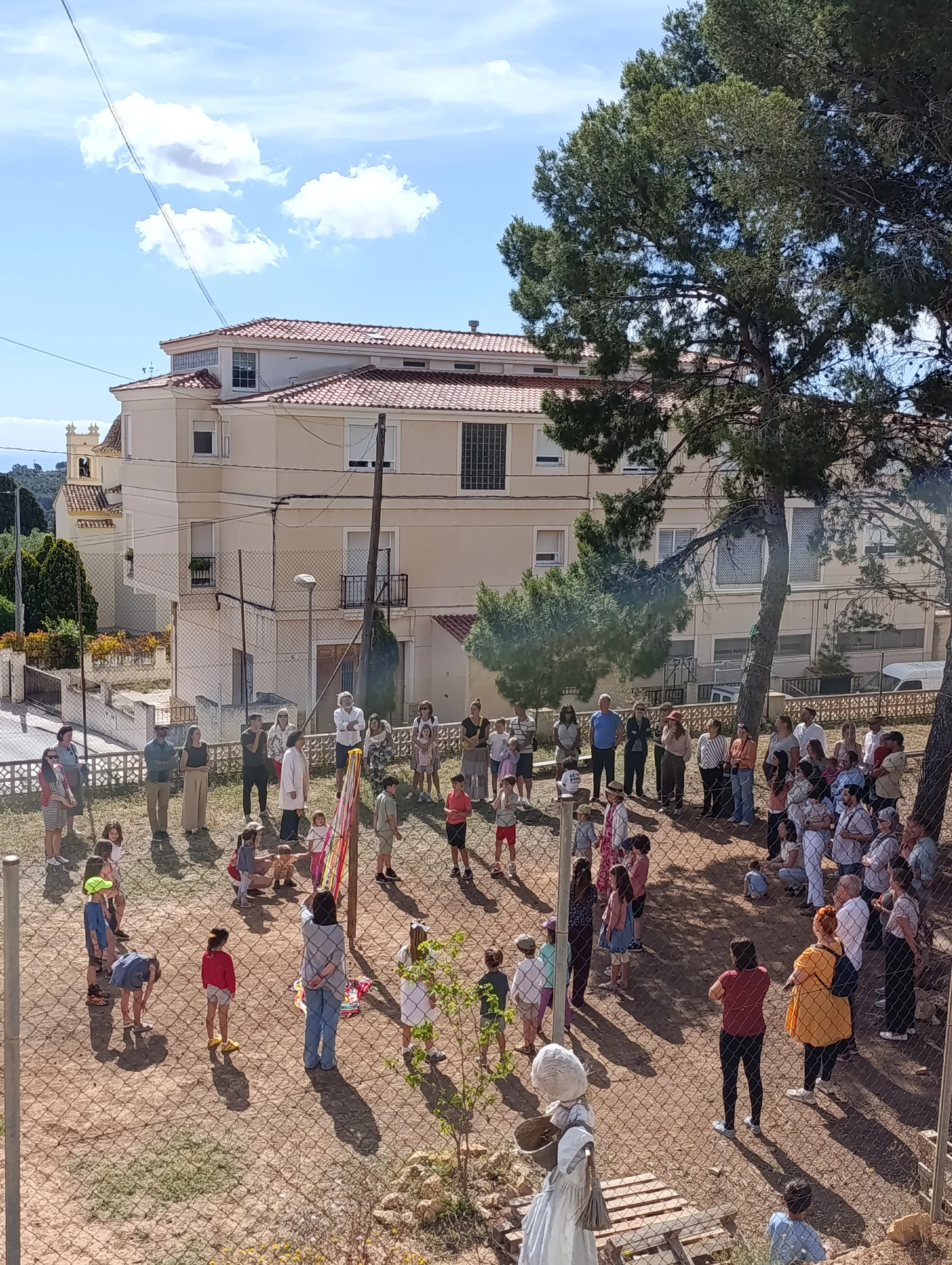 Grupo de personas reunidas en un espacio al aire libre, algunas de ellas sosteniendo banderas, frente a una cerca de alambre y con edificios y árboles al fondo en un día soleado.