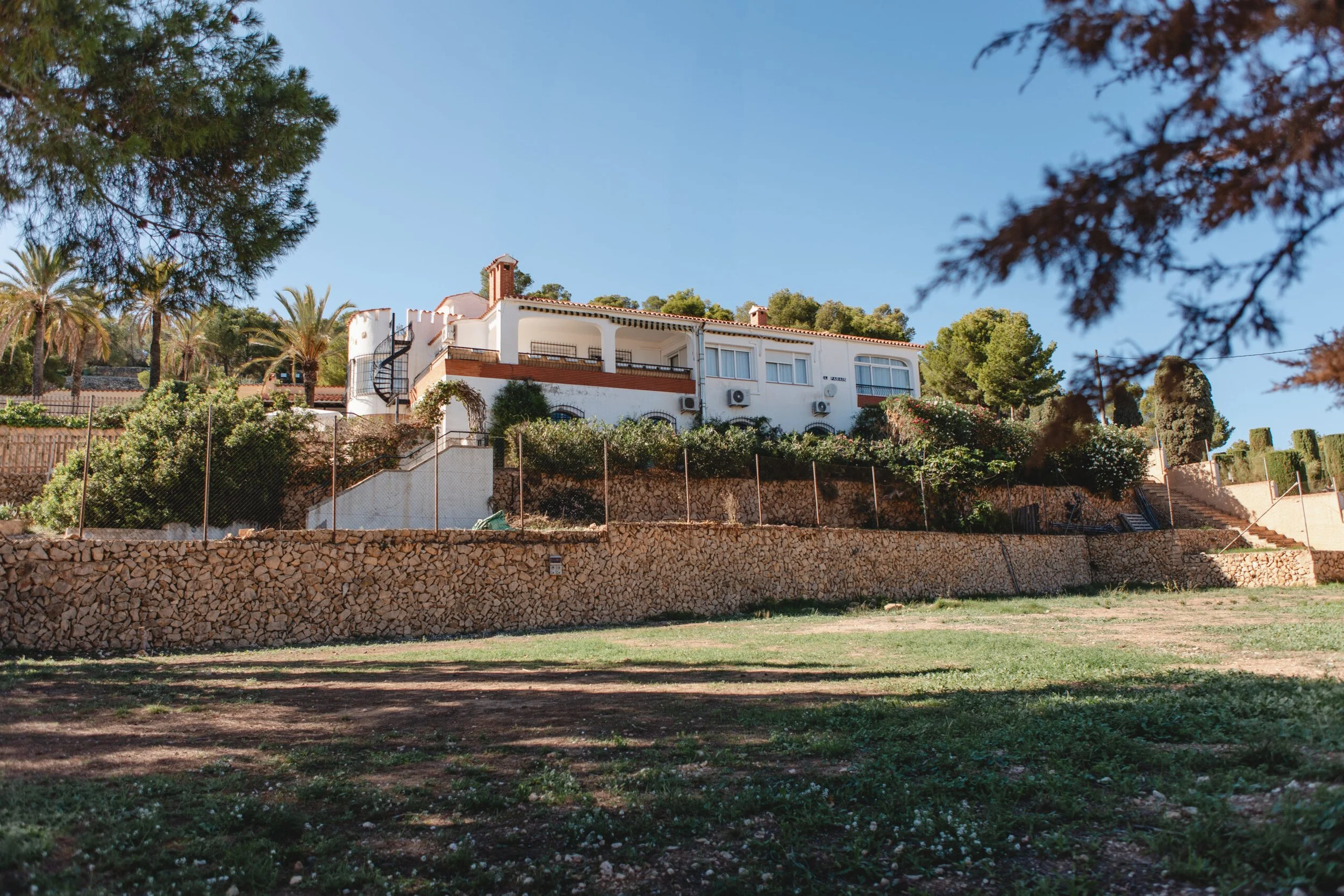 Casa blanca de dos pisos con balcones y aire acondicionado en el exterior, rodeada de palmeras y árboles, con un muro de piedra en primer plano y un camino de escalones a la derecha.