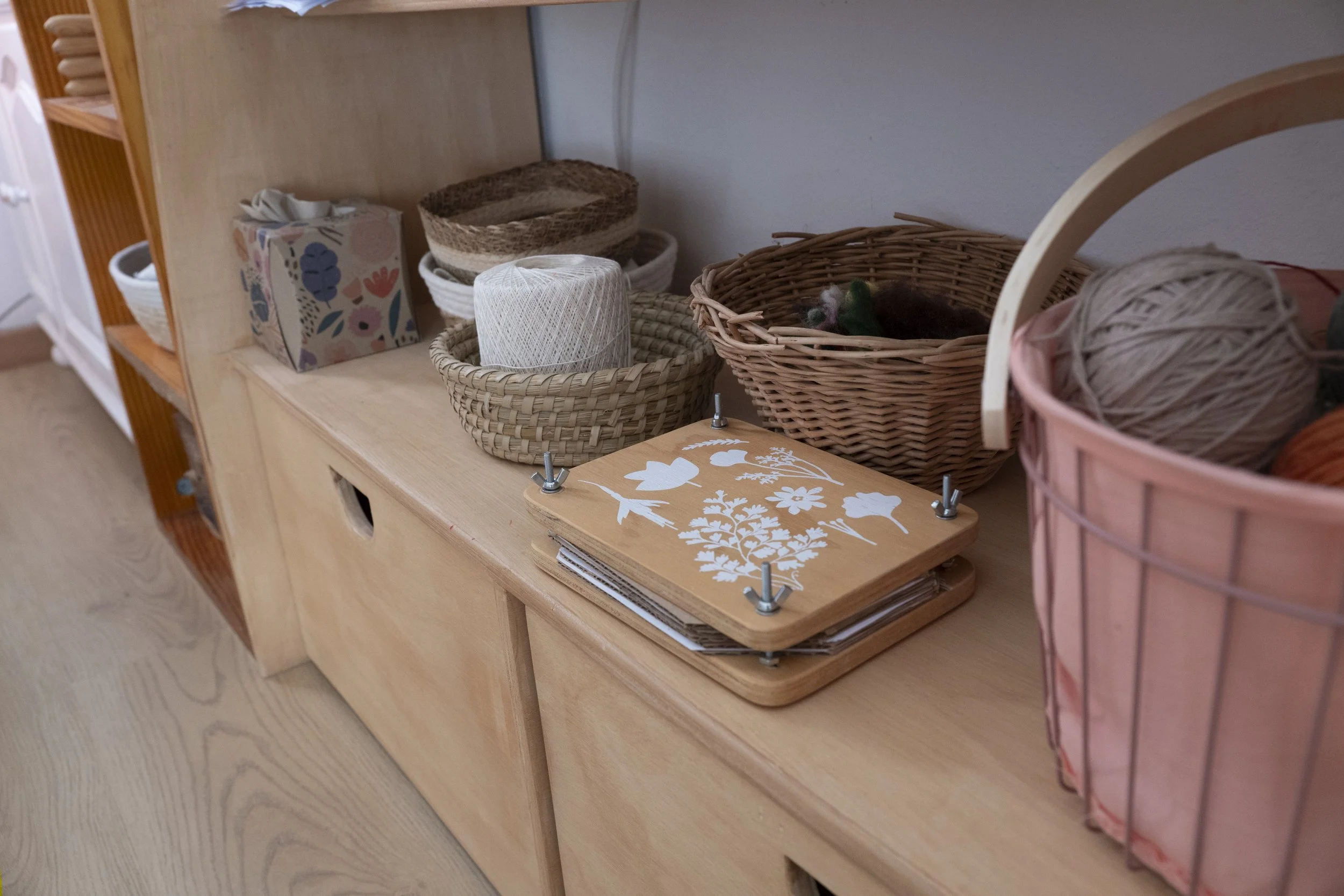 Estantes de madera con cestas de cuerda y una caja de cartón decorada con flores y pájaros, junto a una caja de hilos de lana y un pequeño gato escondido en una de las cestas.
