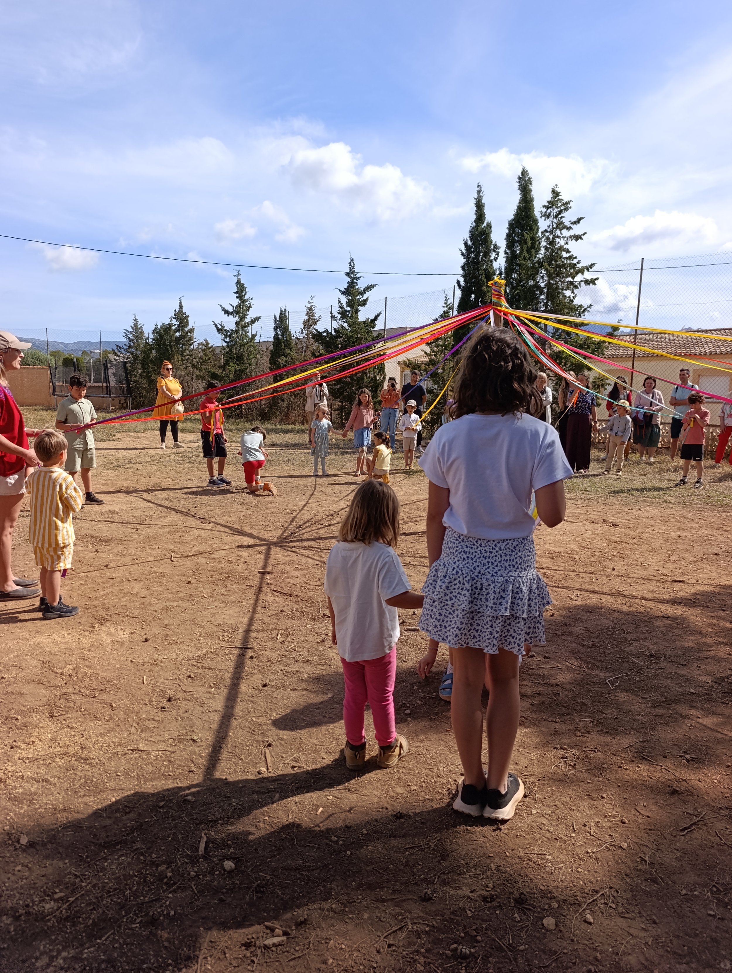 Niños y adultos participando en una actividad con una piñata con ritmo en un espacio abierto con árboles y un muro de malla, en un día soleado.