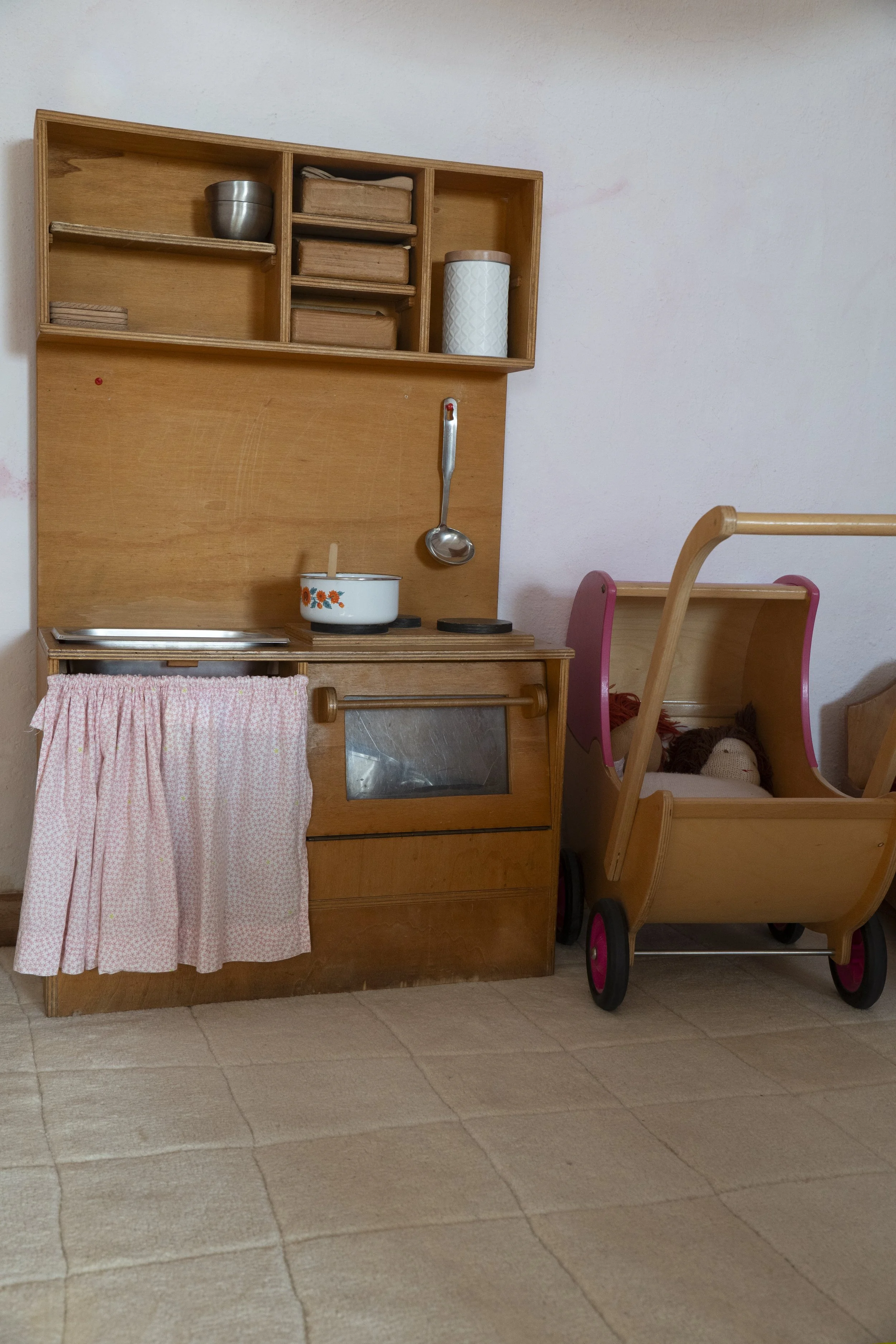 cocina de juguete de madera con estantes, ollas y utensilios, cochecito de bebé de madera con ruedas rosas y un peluche