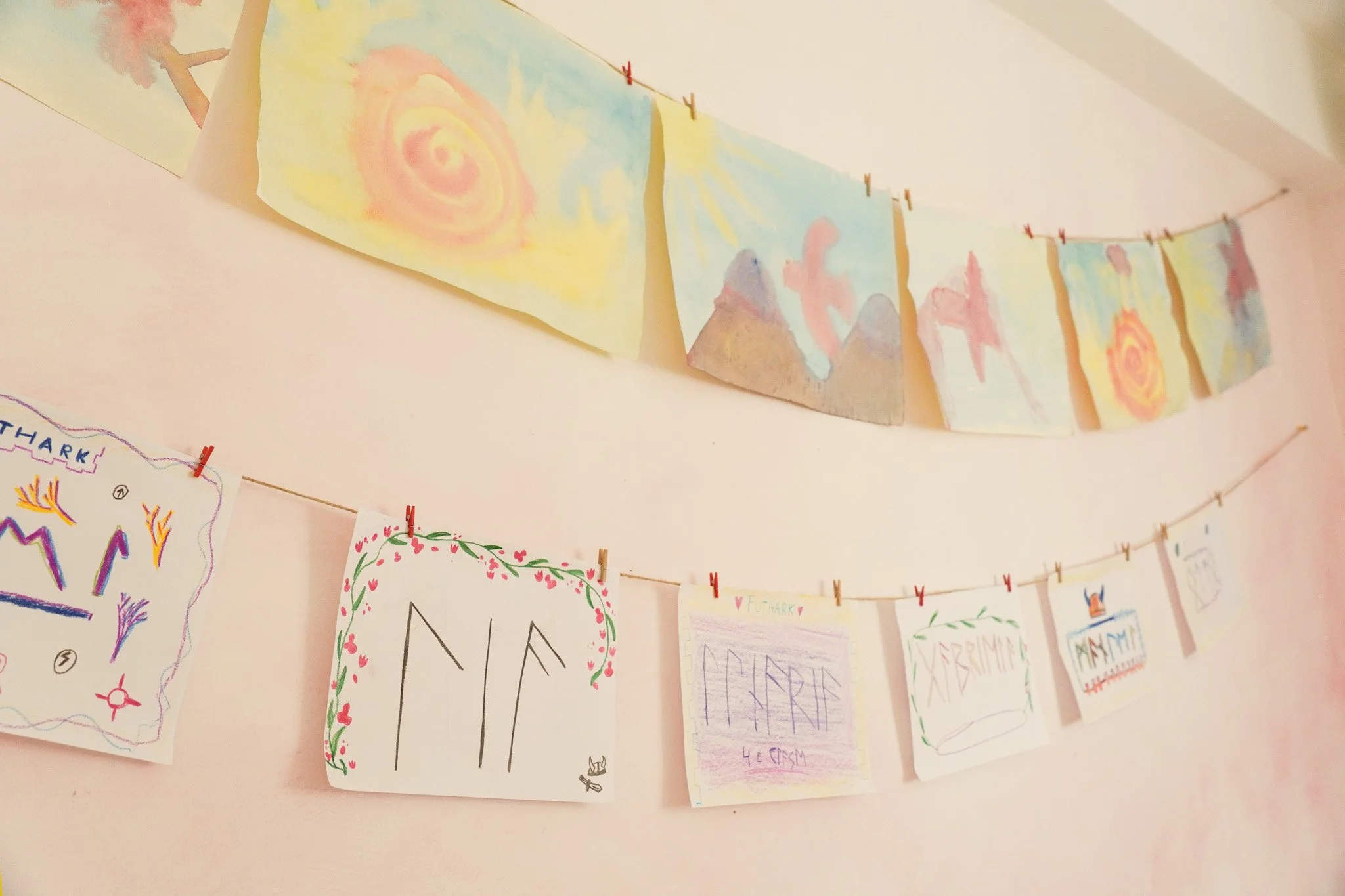 Tarjetas de arte hechas a mano colgadas con pinzas rojas y verdes en una cuerda, decoradas con dibujos y mensajes coloridos, en un fondo de pared de color rosa claro.