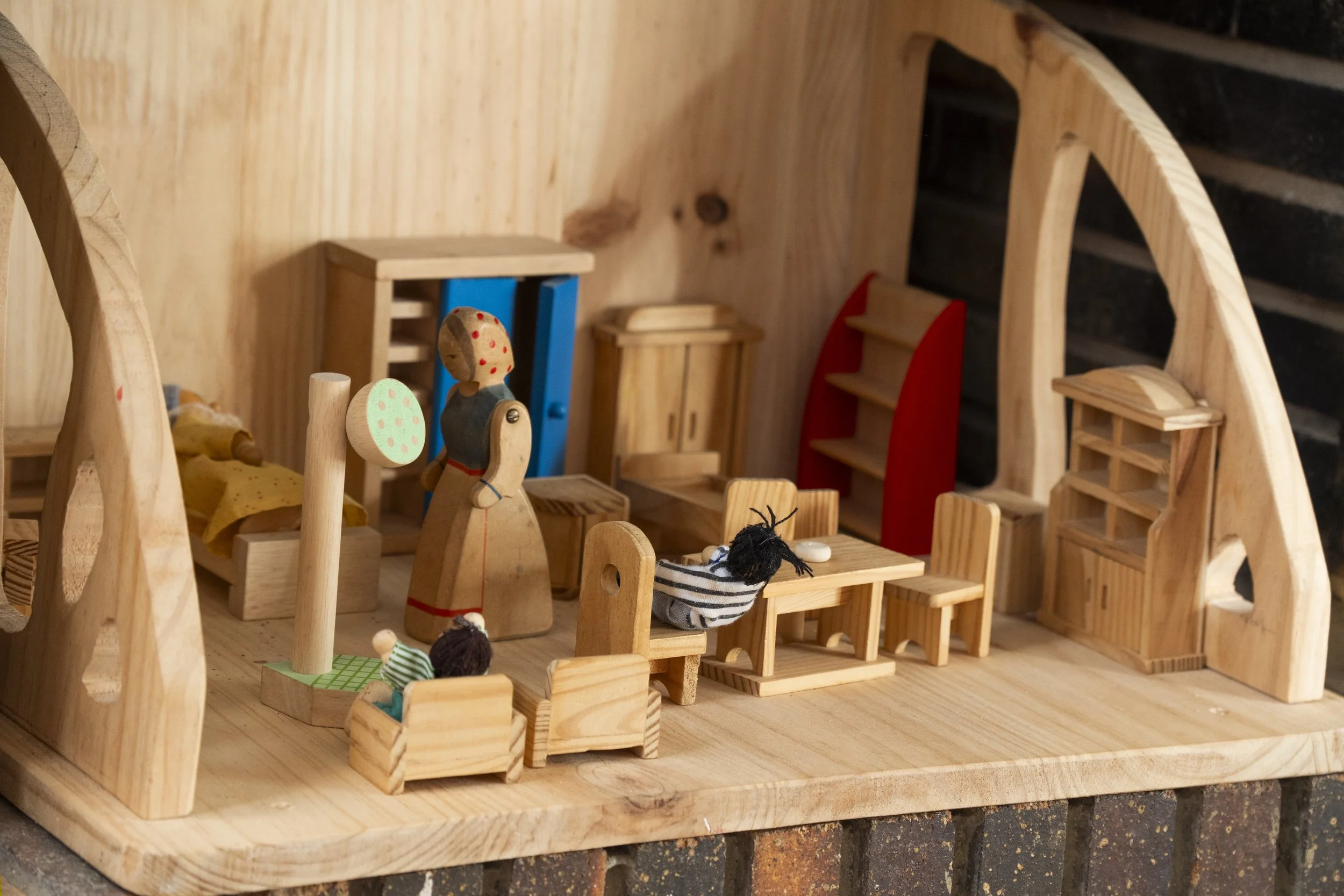 Escena interior de una construcción de madera con muebles pequeños y figuras de muñecos de madera, incluyendo una cama, estantes y sillas, en un ambiente que parece una maqueta de una casa.